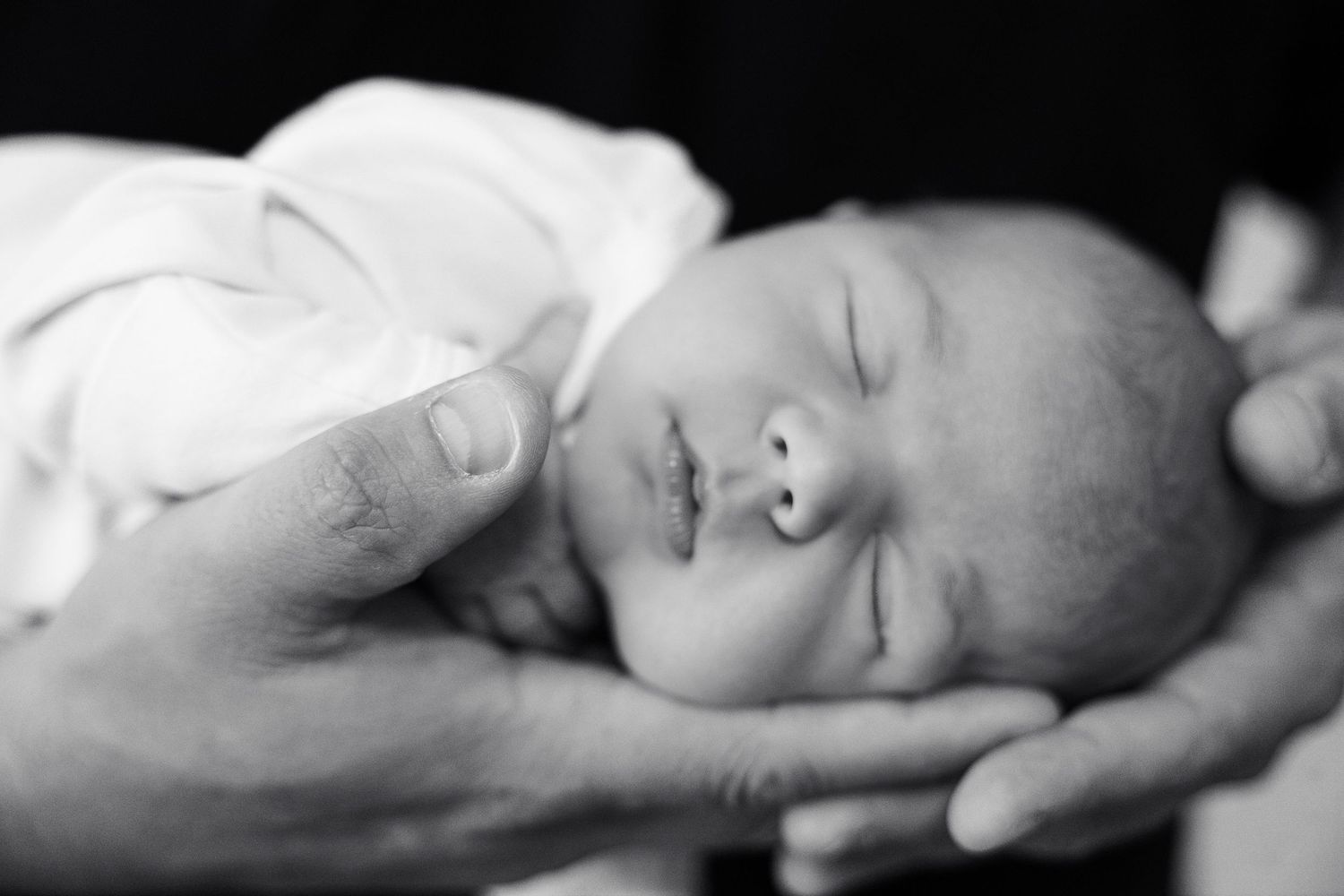 Black and white photo of a sleeping newborn baby cradled gently in adult hands, wrapped in a white blanket.