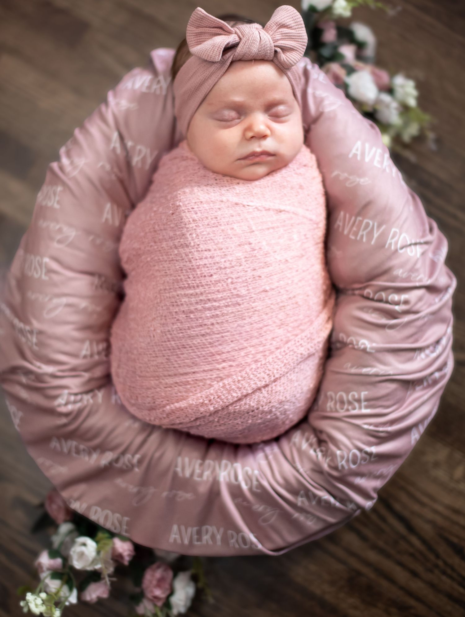 Newborn baby girl swaddled in pink wrap with bow headband, resting in a round pillow printed with Avery Rose.