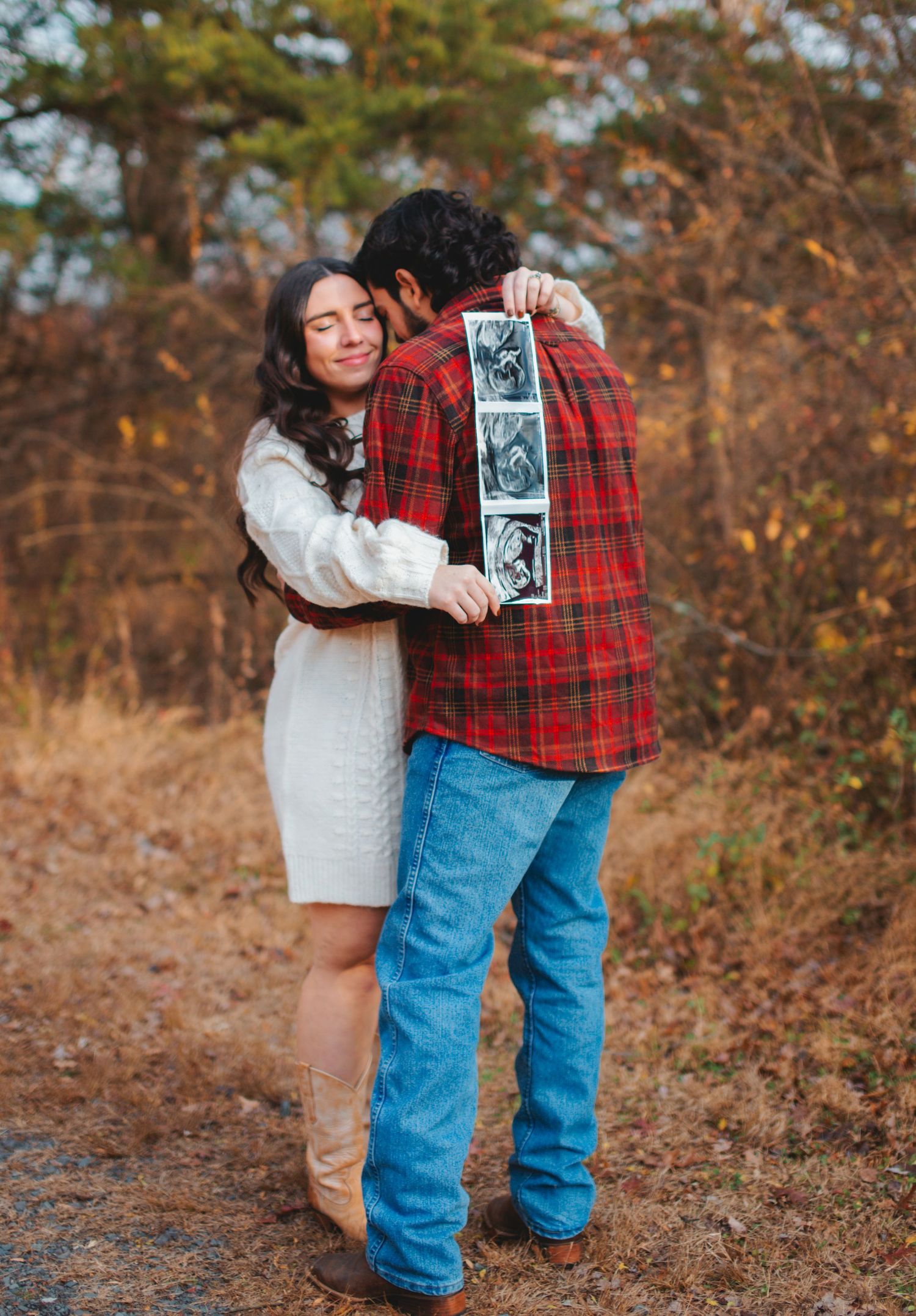 A couple holds up an ultrasound photo while embracing in a wooded autumn setting wearing cozy sweaters and jeans.