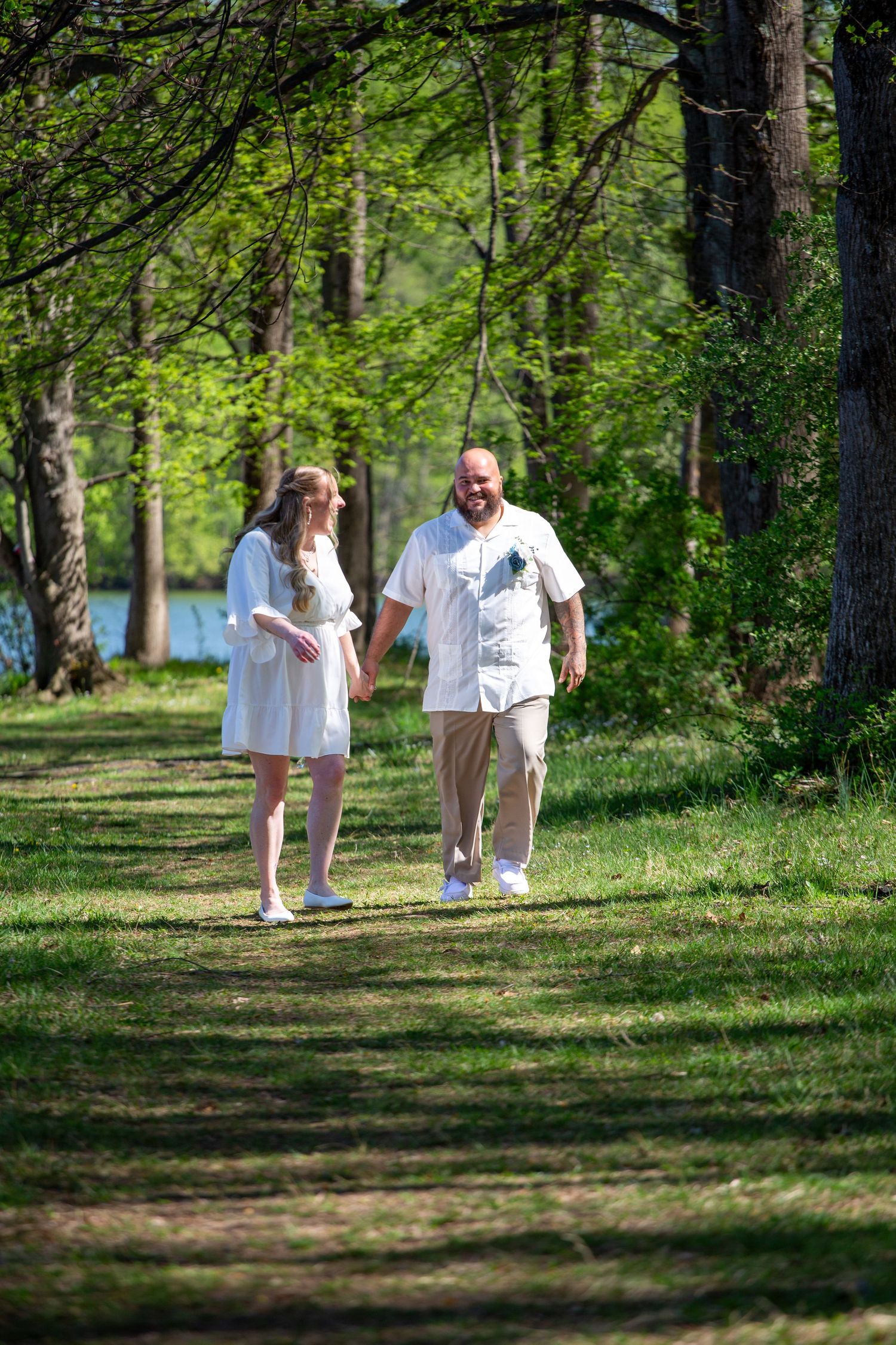 A couple in white attire walks hand in hand along a sunlit forest path surrounded by lush green trees.