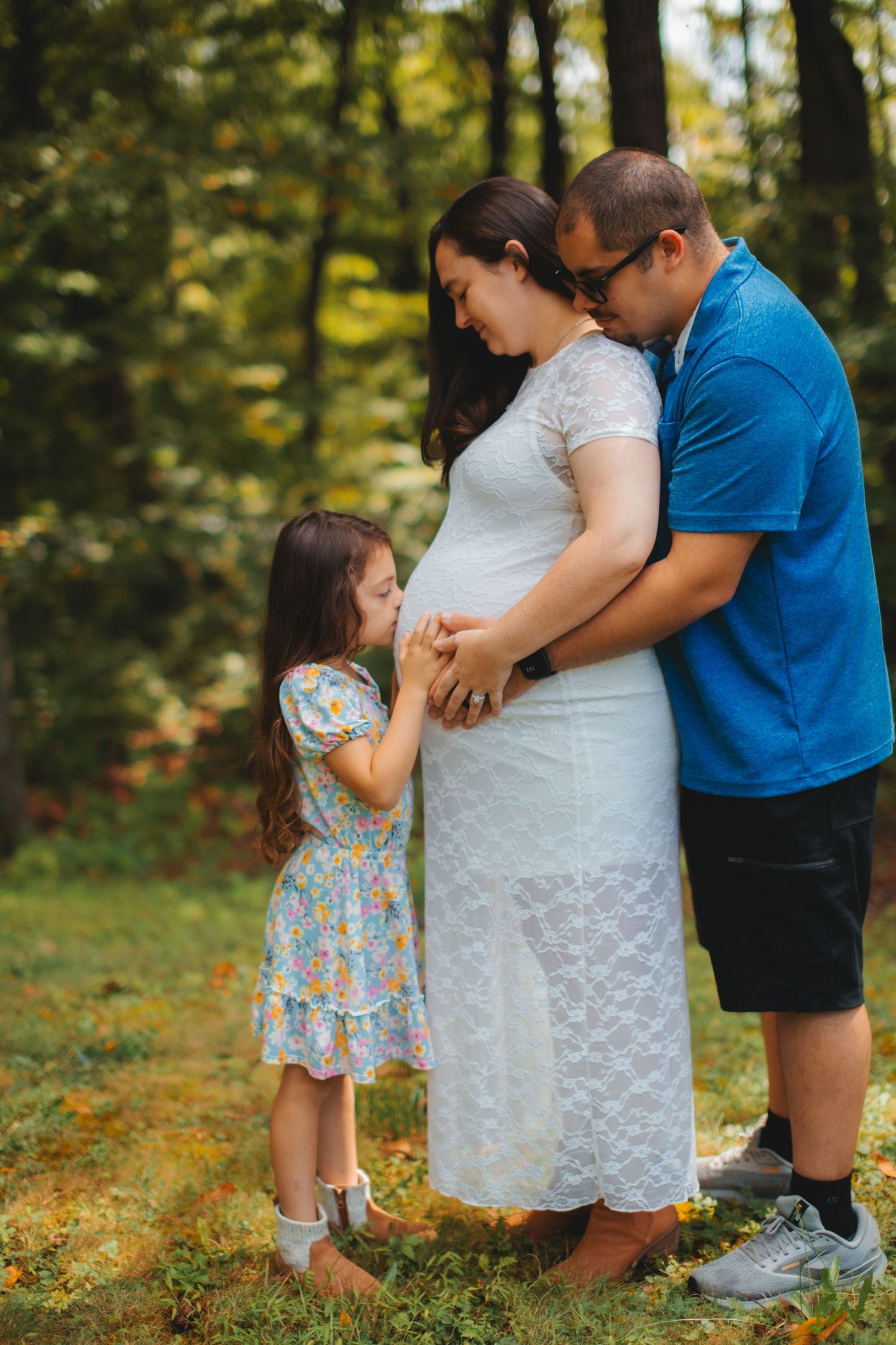 A family in a forest setting shares a tender moment as they await their new arrival, with a young girl in a floral dress.