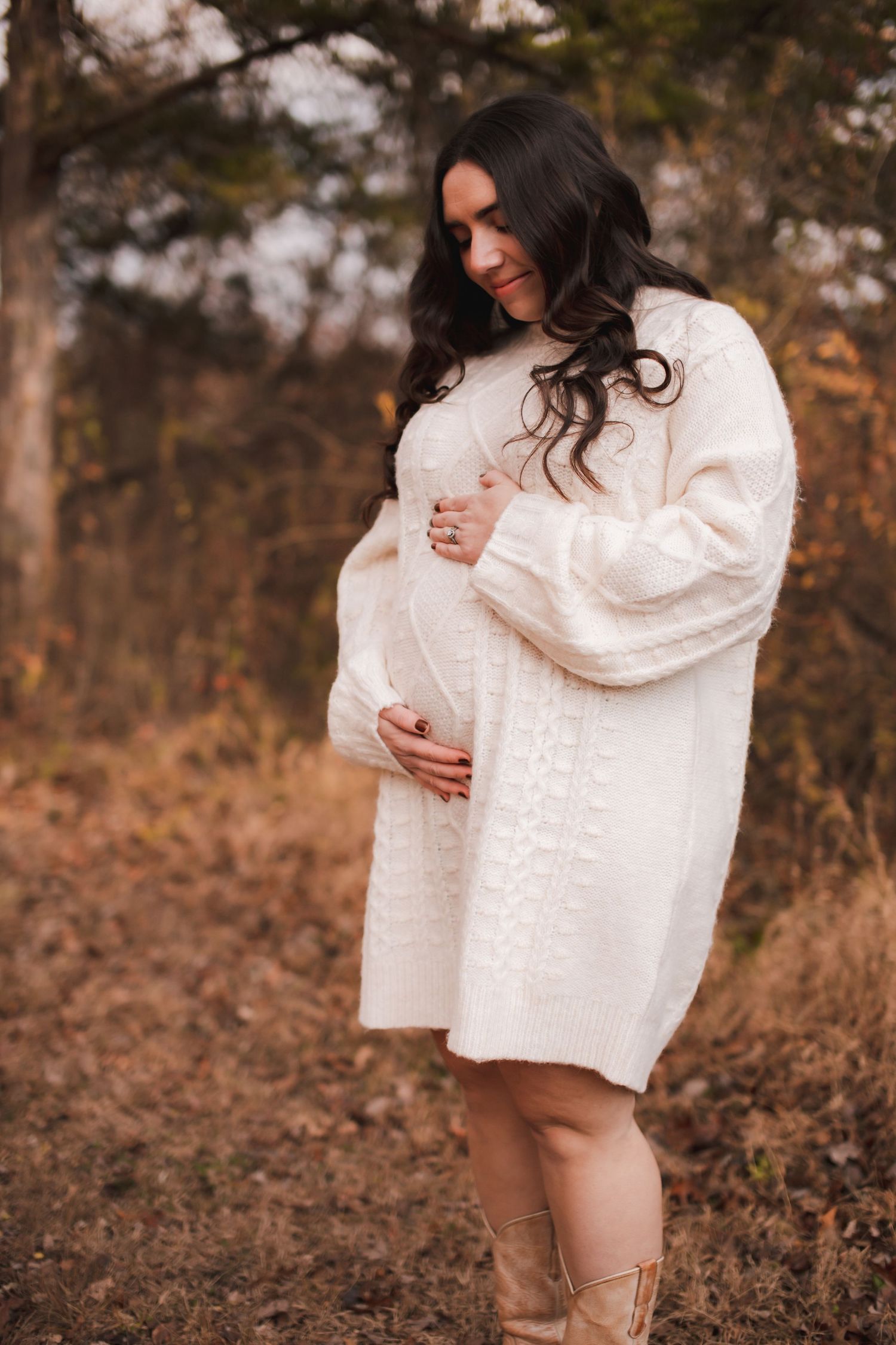 A figure in a cozy white sweater poses against an autumn backdrop with brown foliage and natural lighting.