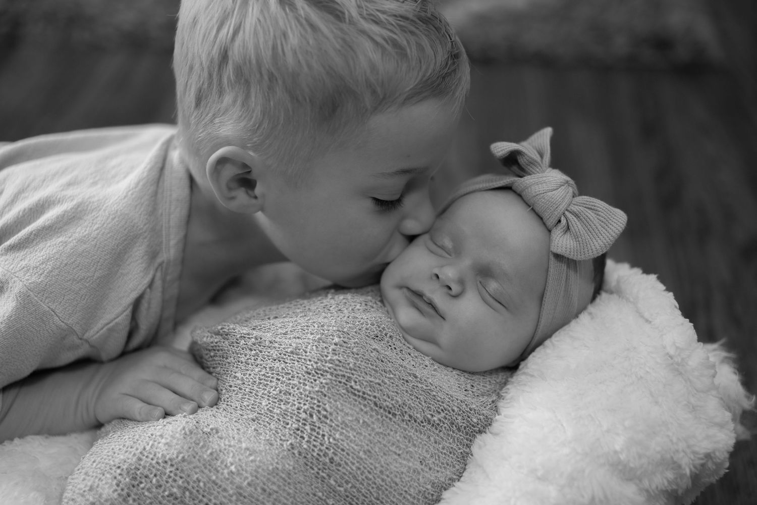 Black and white photo of a young boy kissing his sleeping newborn sister wrapped in a knit blanket with a bow headband.