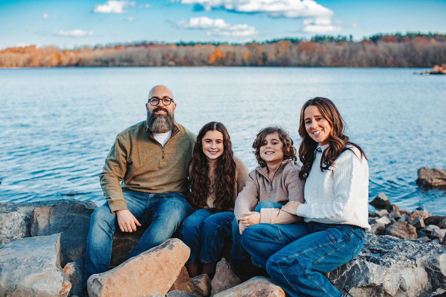 A family relaxes together on large rocks along a scenic lakeshore with autumn trees in the background.