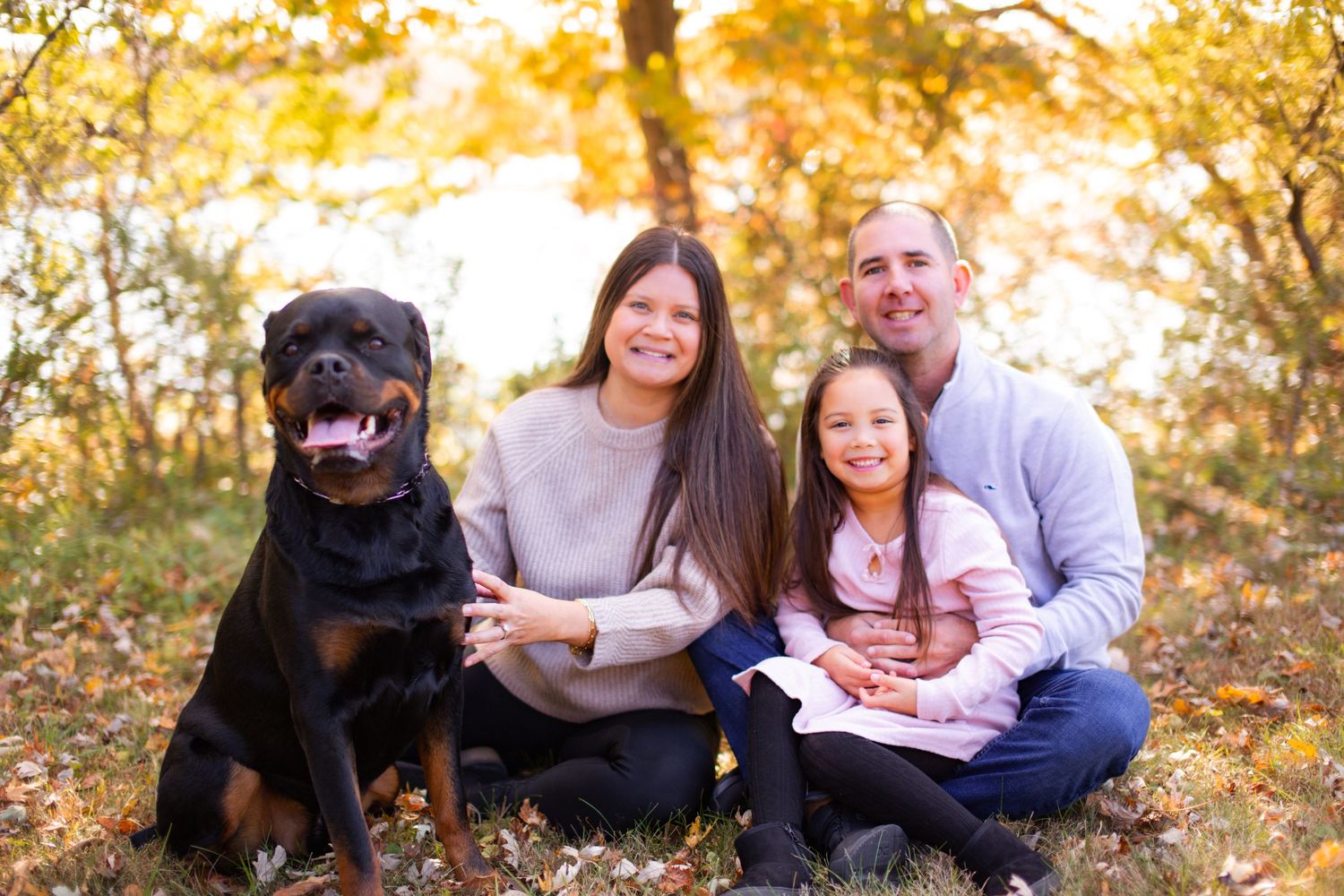 A family portrait with their black Rottweiler dog in a fall setting with golden leaves.