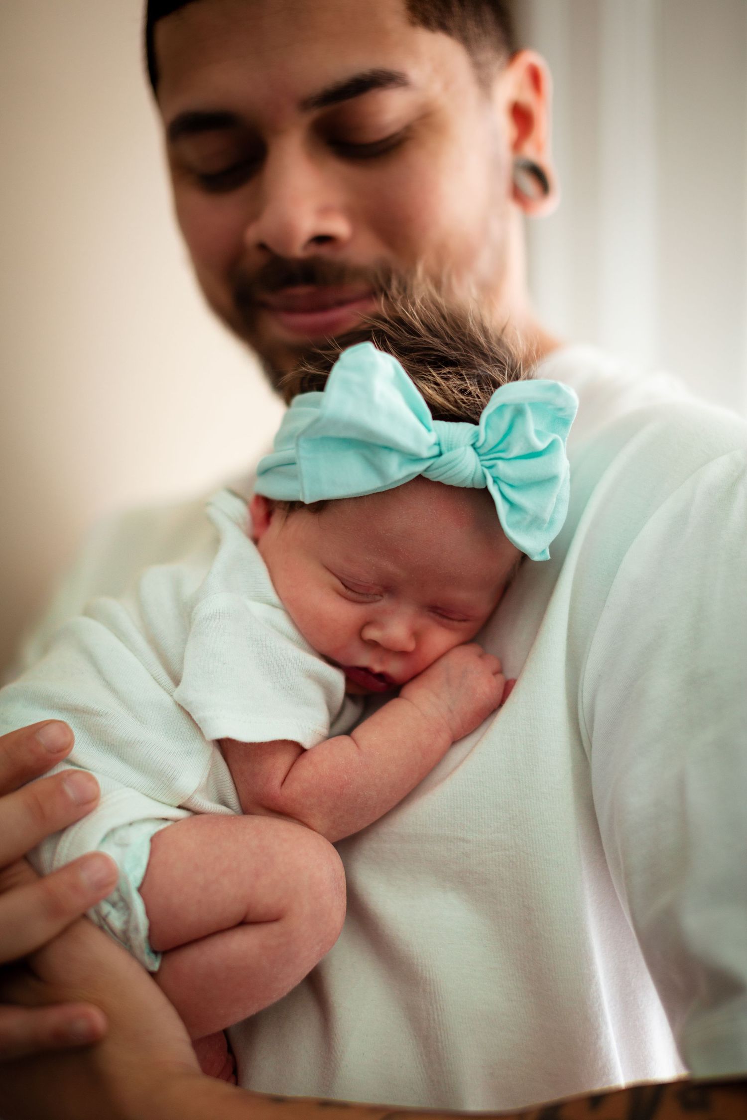 A sleeping newborn baby wearing a mint green bow headband rests peacefully against someone's chest.