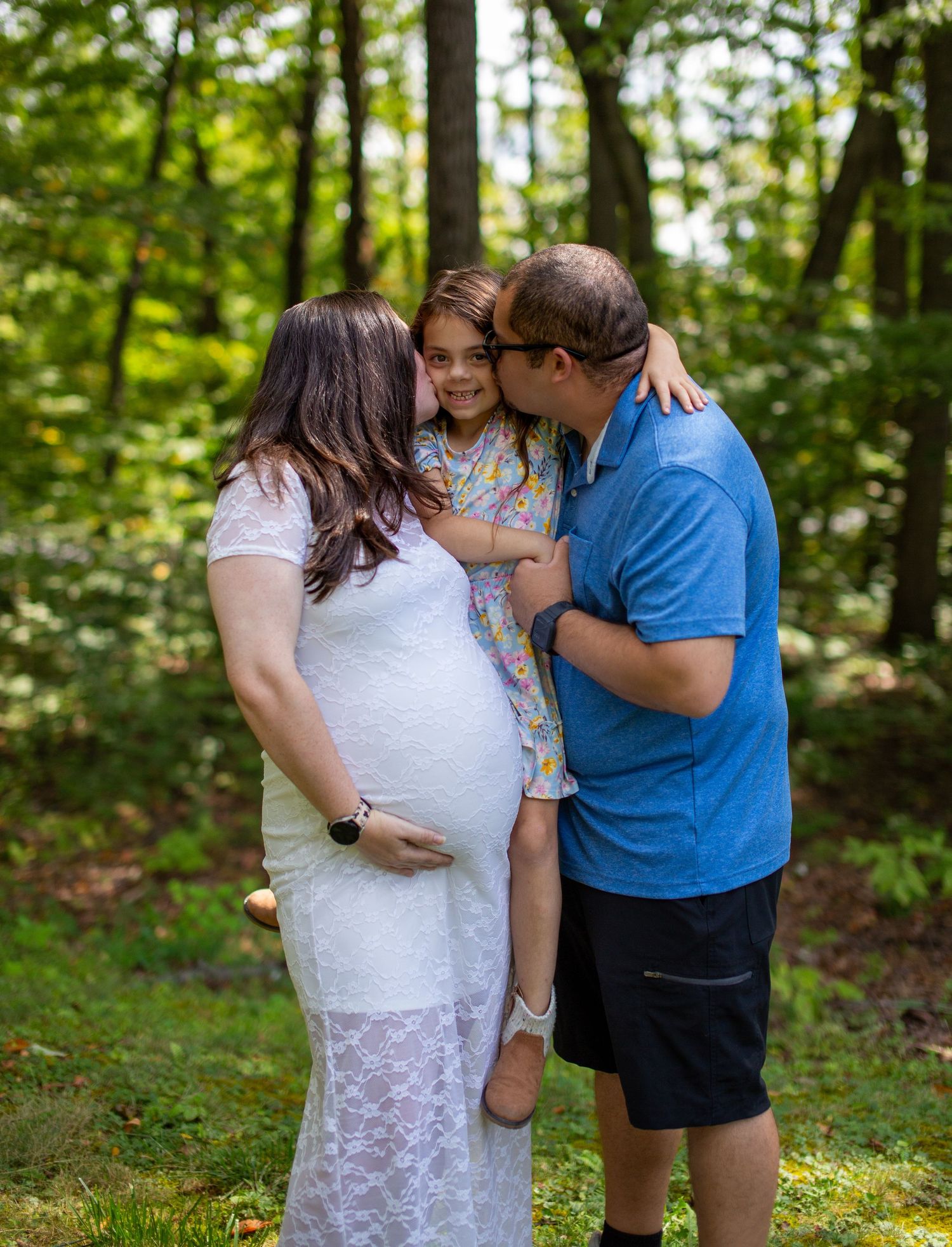 Family maternity photos taken outdoors in a sunlit wooded setting.