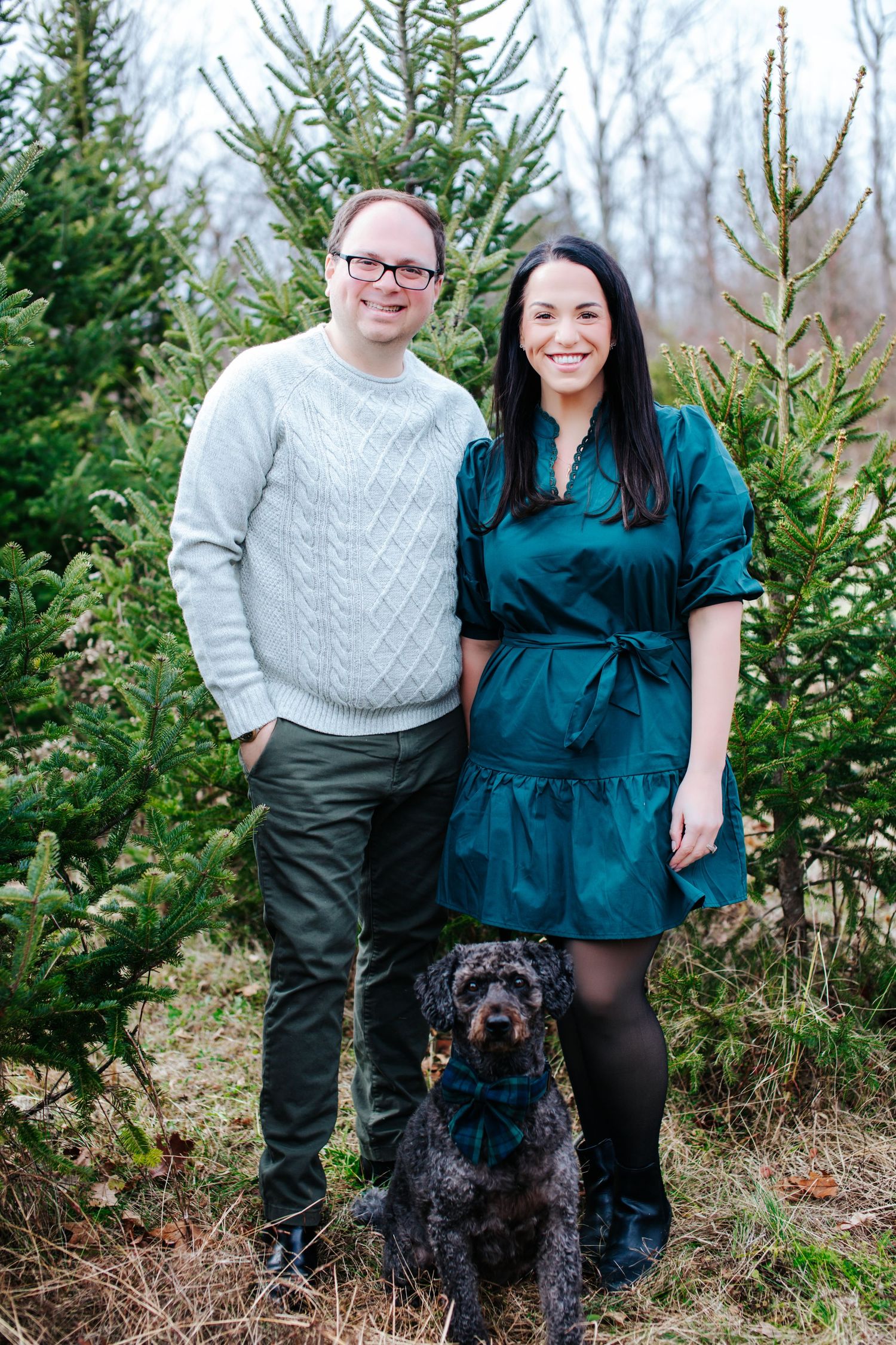 A couple in winter attire poses with their black dog in front of evergreen trees.