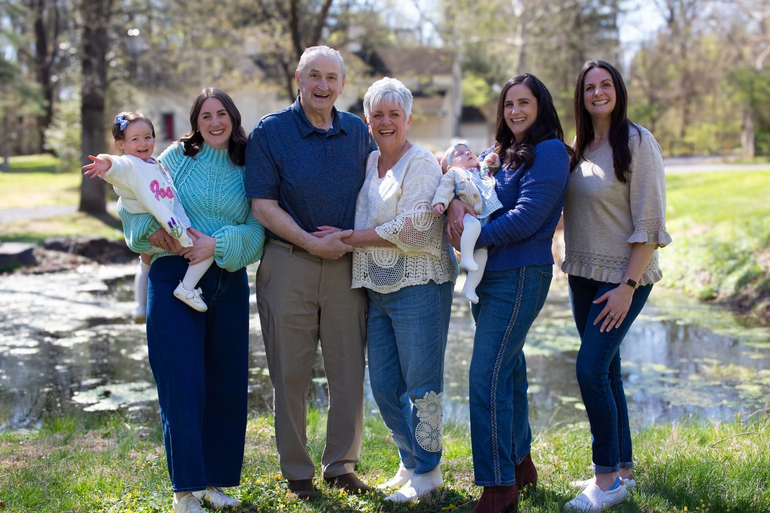 Multi-generational family of six poses outdoors near a pond, with two women holding babies flanking an older couple.