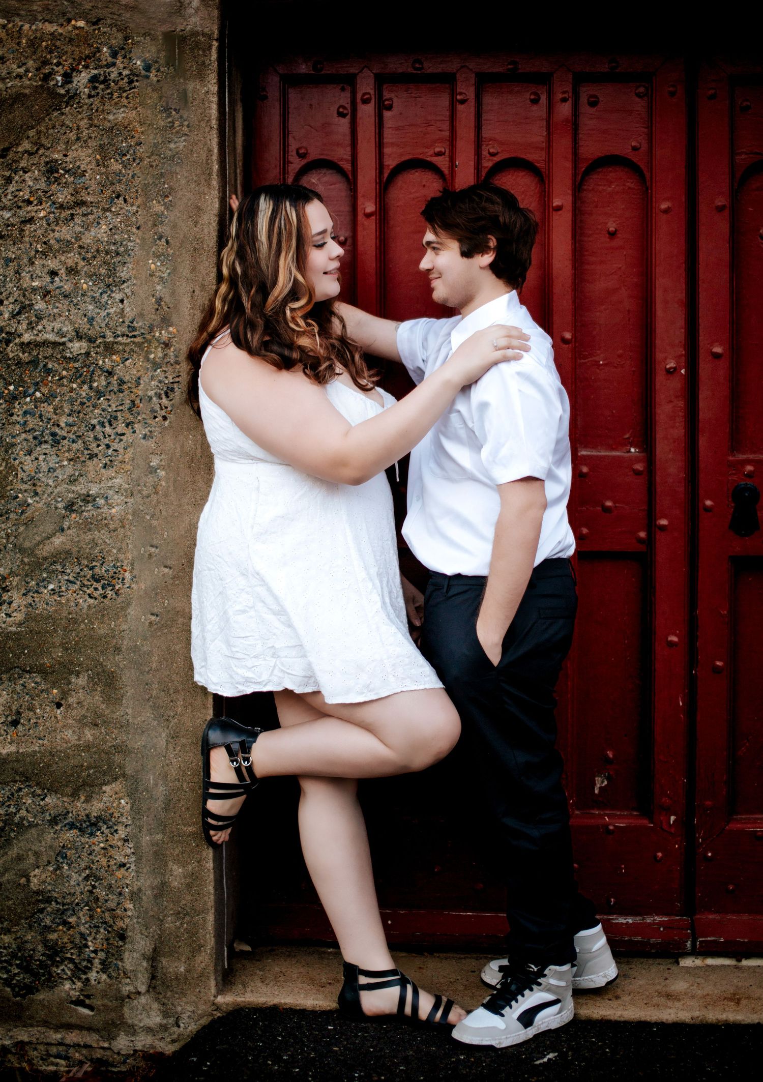 A playful couple poses against red doors in casual white outfits.