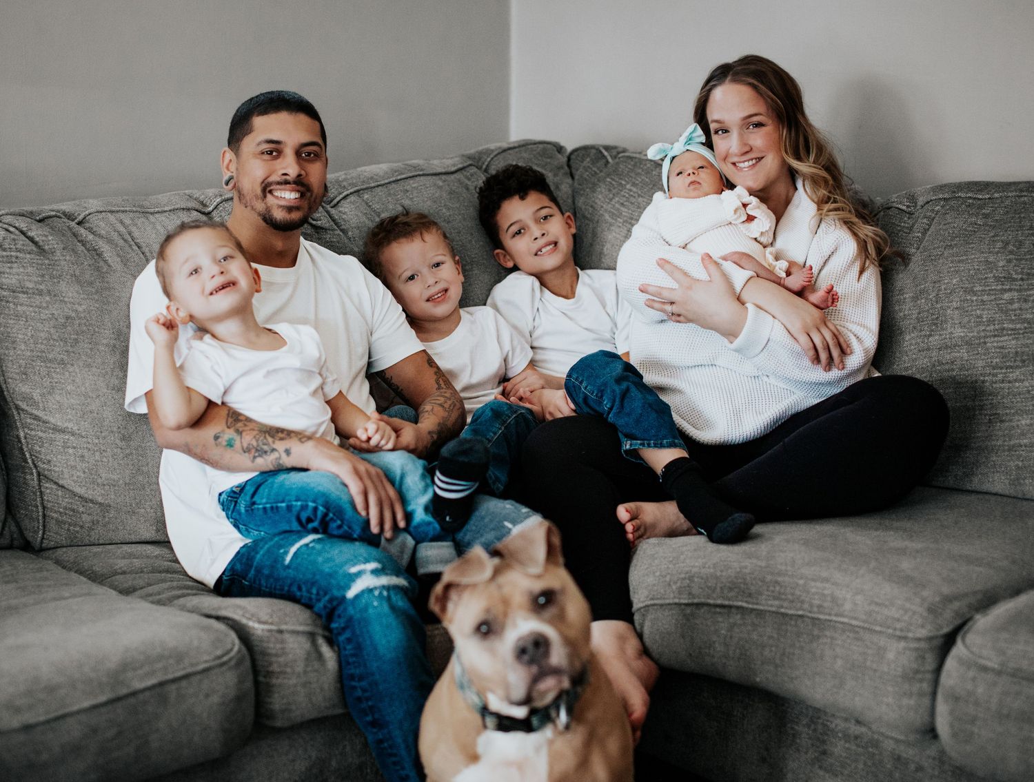 Happy family with three boys and a newborn baby posing on a gray couch with their brown dog.