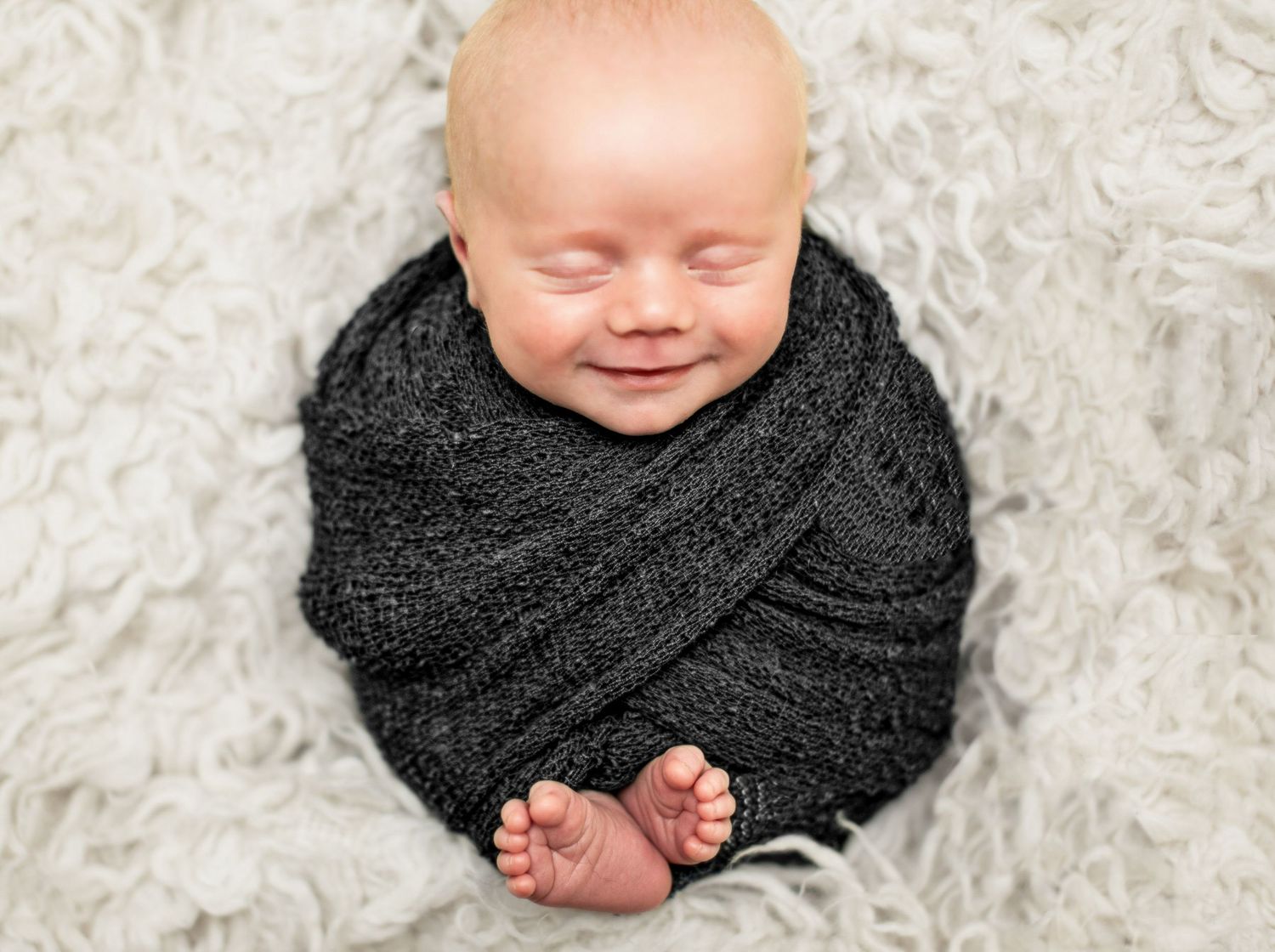 Baby wrapped in black knit blanket on white fluffy carpet.