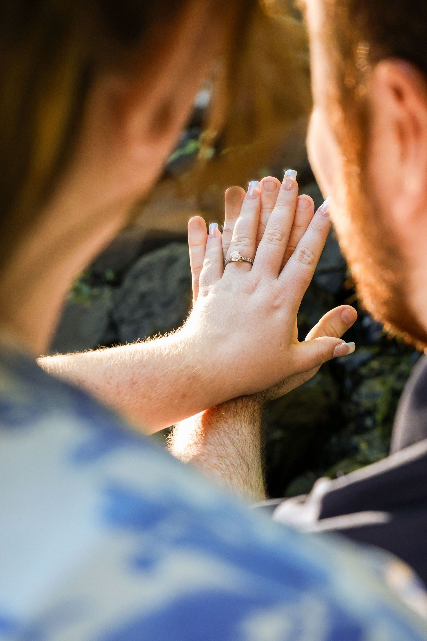 Couple comparing hands outdoors, woman wearing a diamond engagement ring, backlit by warm golden sunlight.