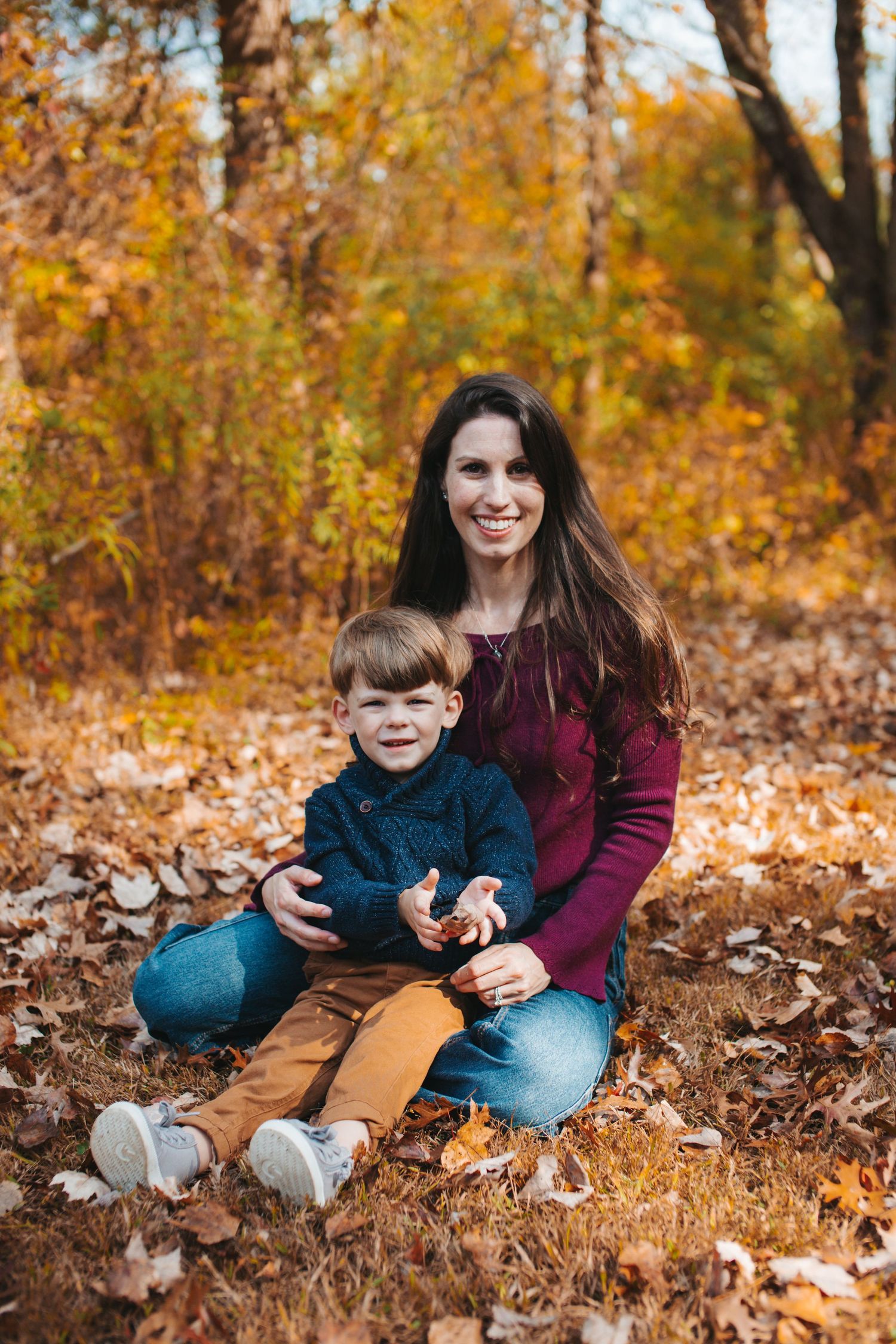 Two people sit together in fallen autumn leaves during a fall photo session in a forest setting.