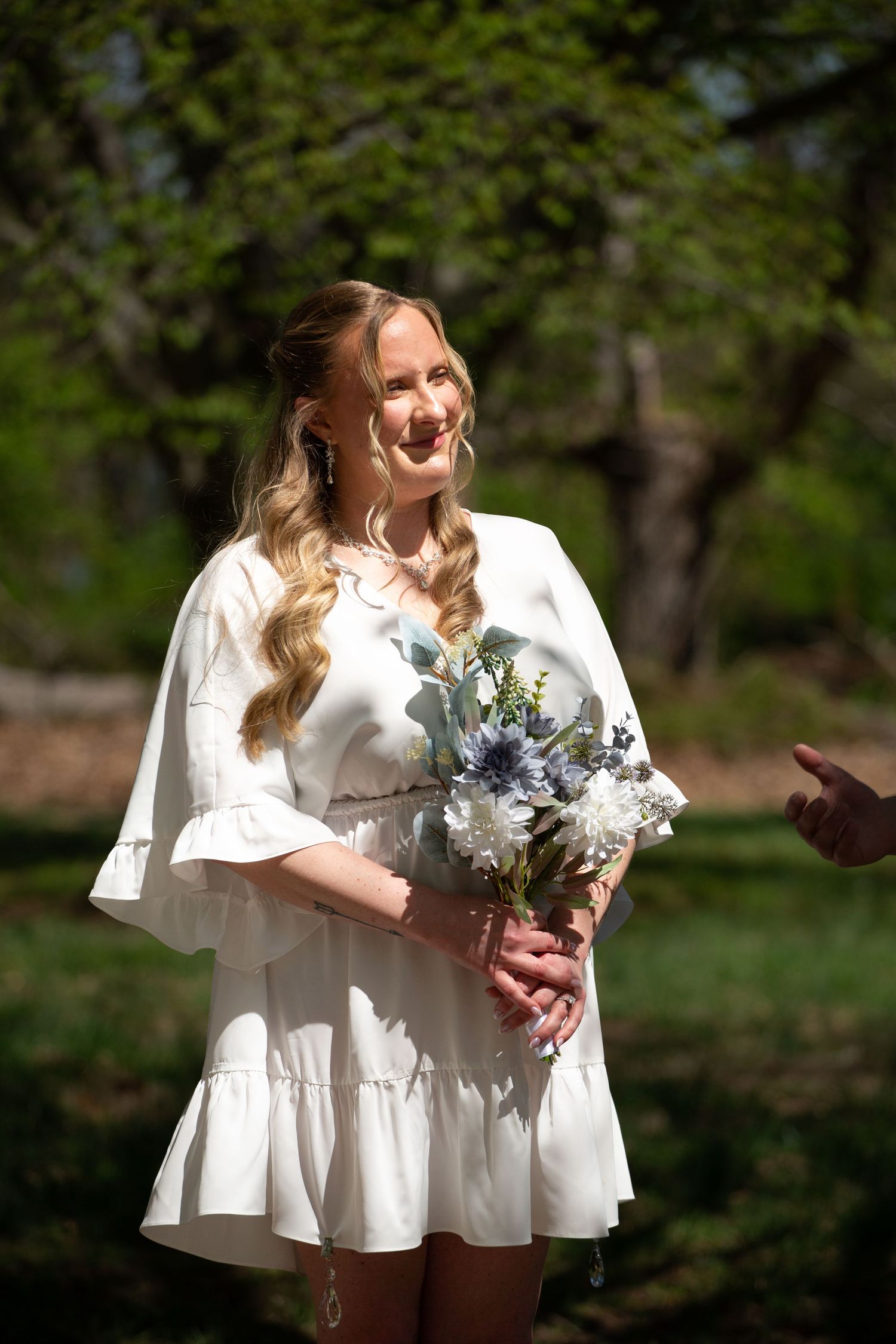 Smiling blonde bride in white dress holding blue and white bouquet outdoors in sunlit garden setting.