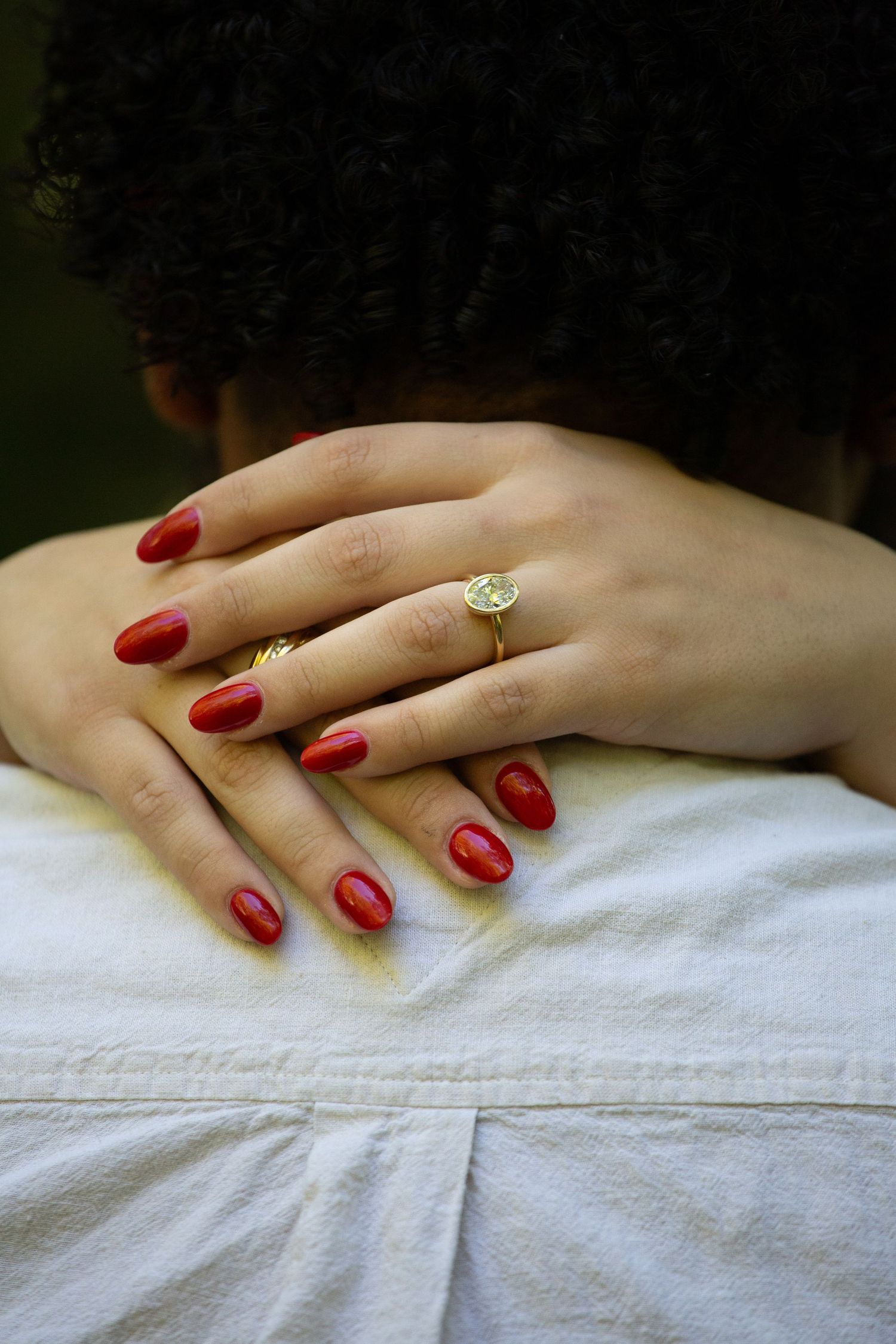 Delicate hands with red nail polish and an engagement ring rest on white fabric.