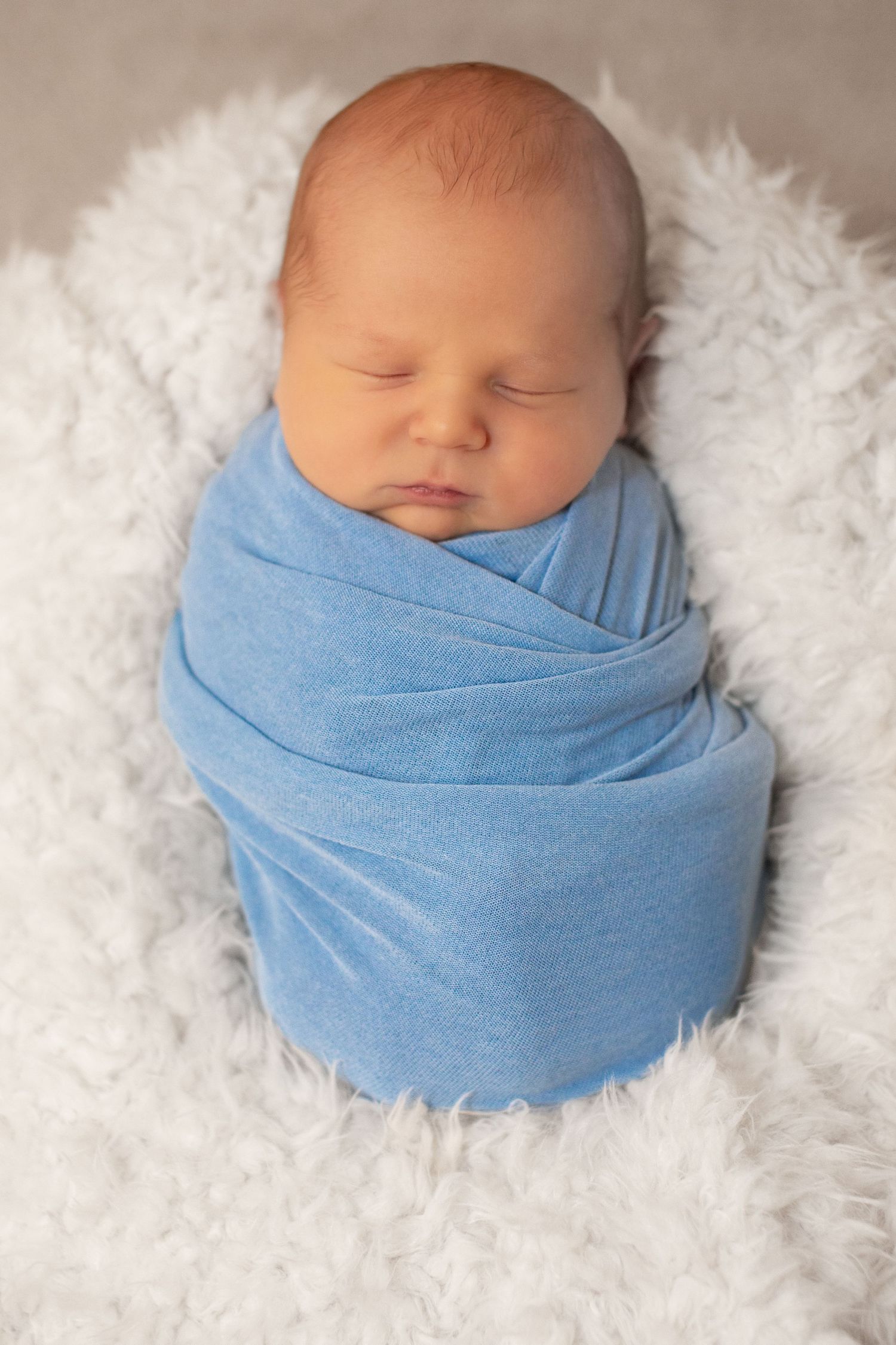 Peaceful newborn baby swaddled in a soft blue wrap, sleeping on a white fluffy blanket.