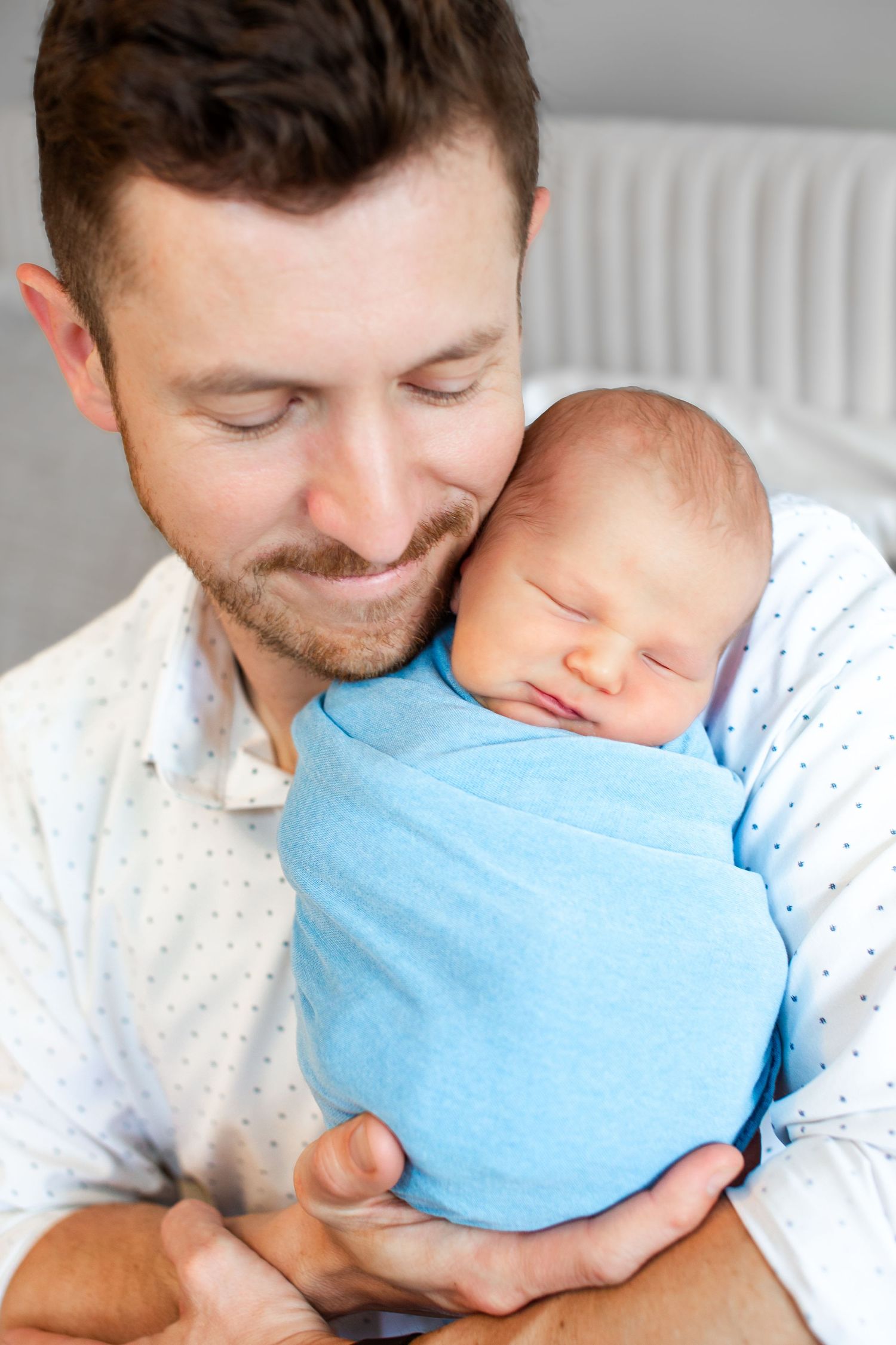 Smiling father with short beard gently holds sleeping newborn baby swaddled in blue blanket close to his cheek.