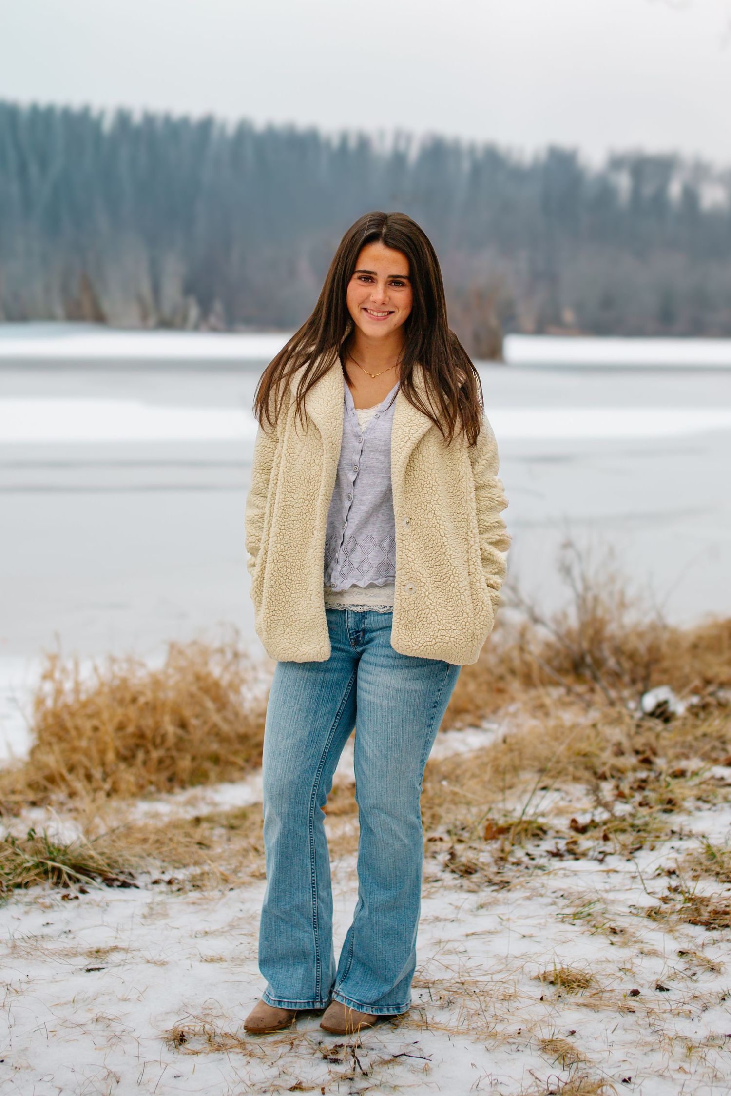 A person in a cream sherpa jacket and jeans poses on a snowy shoreline with a lake and forest in the background.