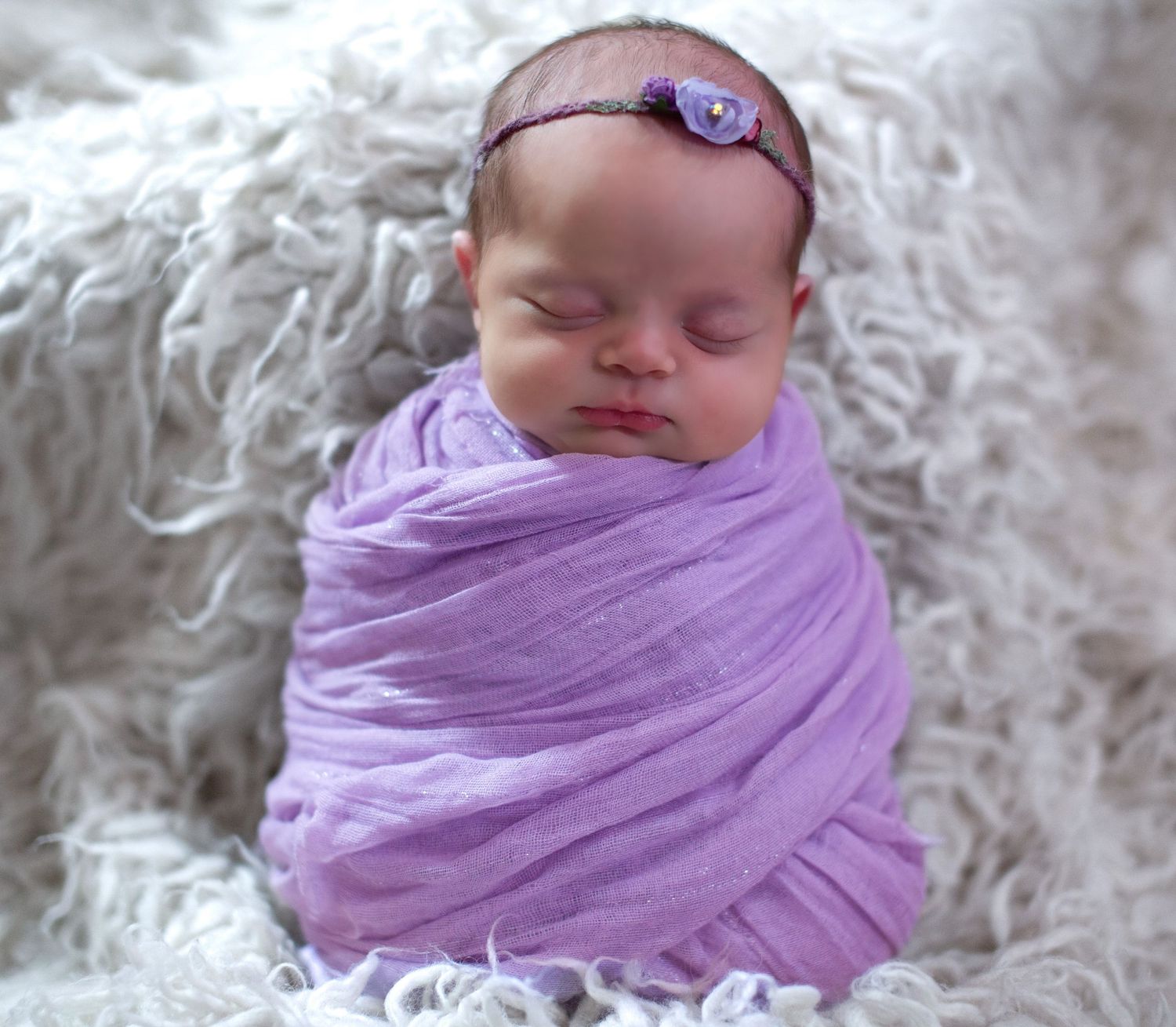 Sleeping newborn wrapped in soft lavender fabric against white textured background.