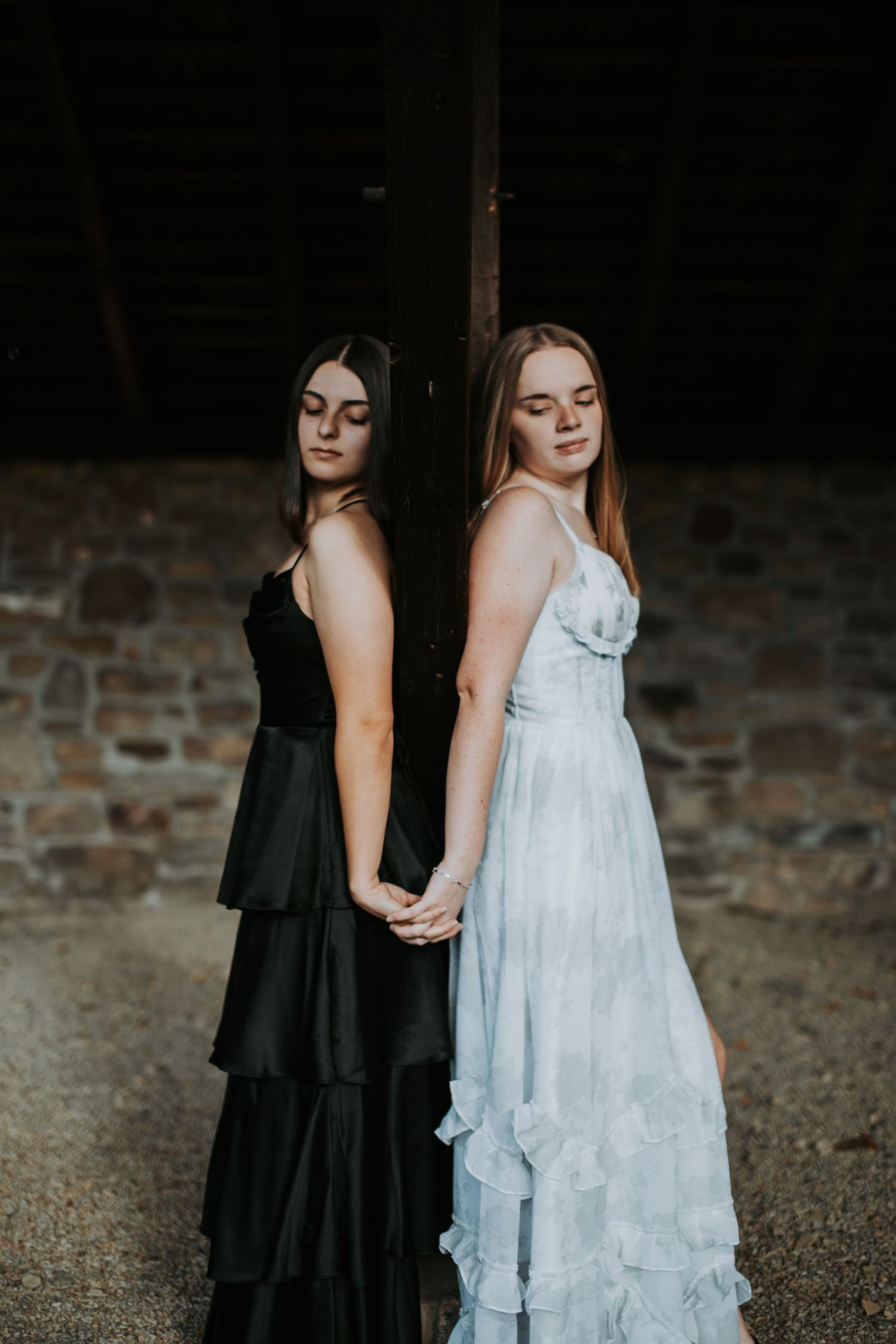 Two people in contrasting black and white dresses pose together against a brick wall in dramatic lighting.