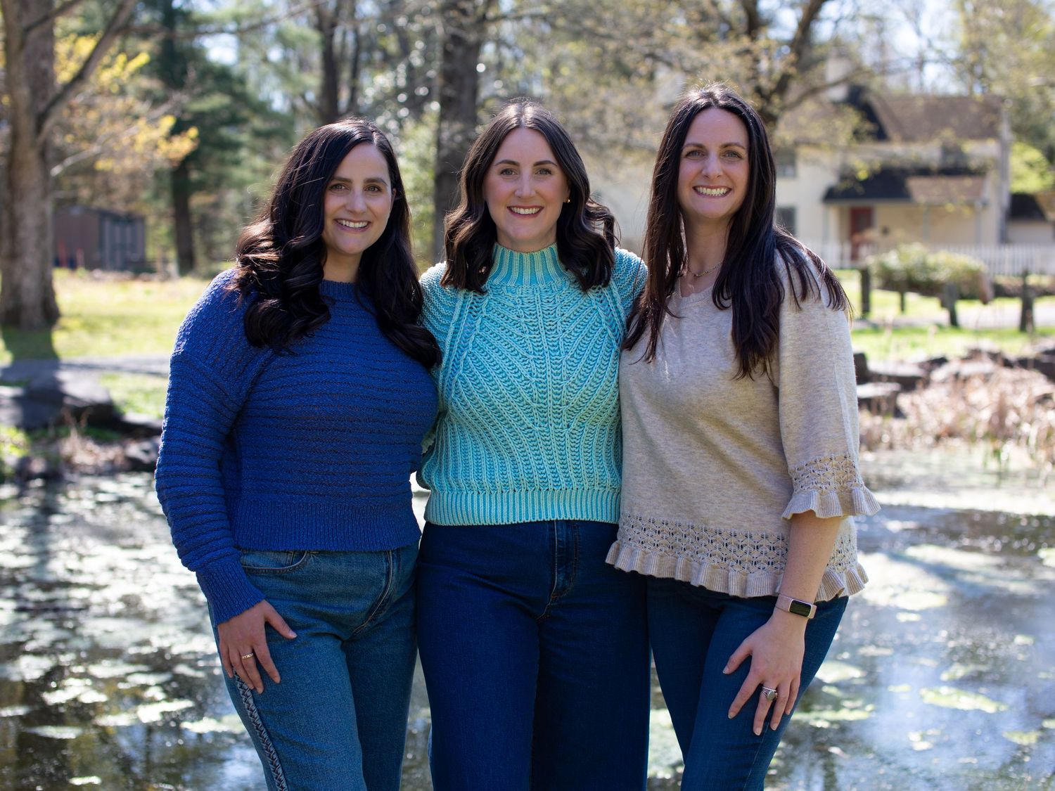 Three smiling women standing together outdoors near a pond in a park on a sunny spring day.