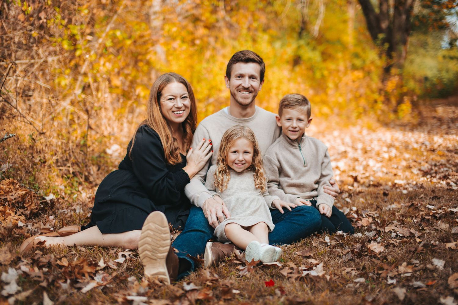 A family sits together on fallen autumn leaves with golden foliage in the background.