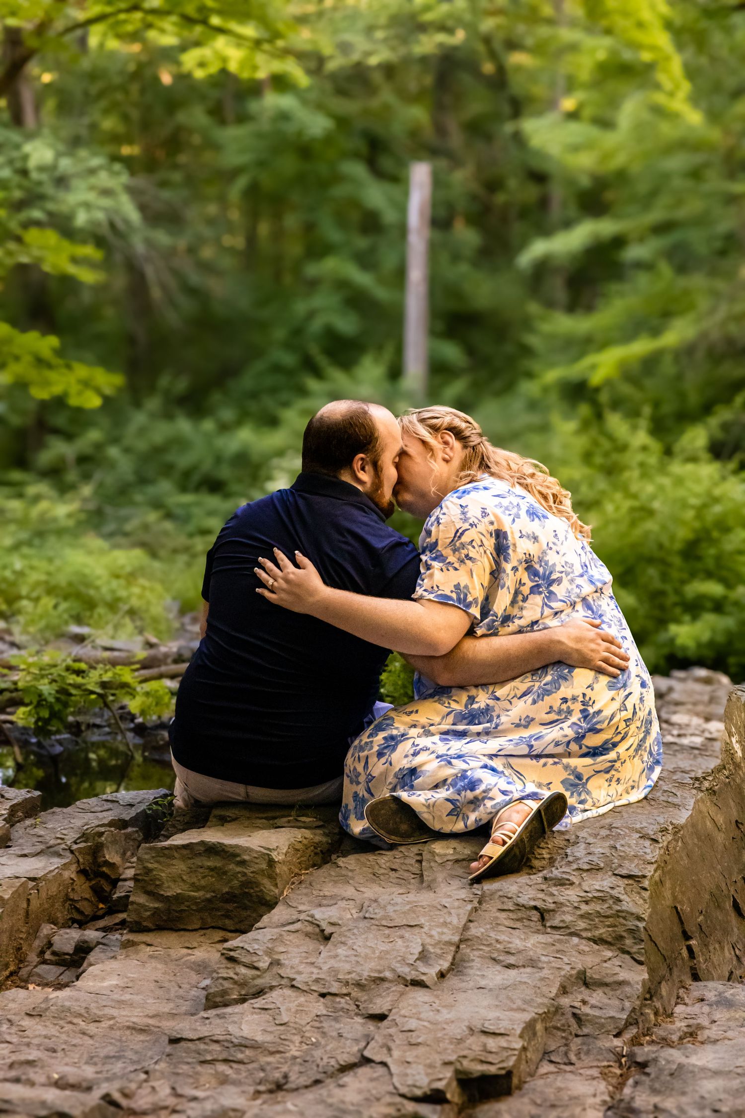 A couple shares a romantic kiss while sitting on rocks surrounded by lush green forest trees.