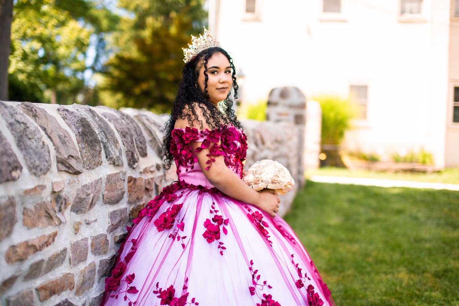 A person in a flowing magenta and white ballgown poses in front of a stone wall during a sunny outdoor photoshoot.