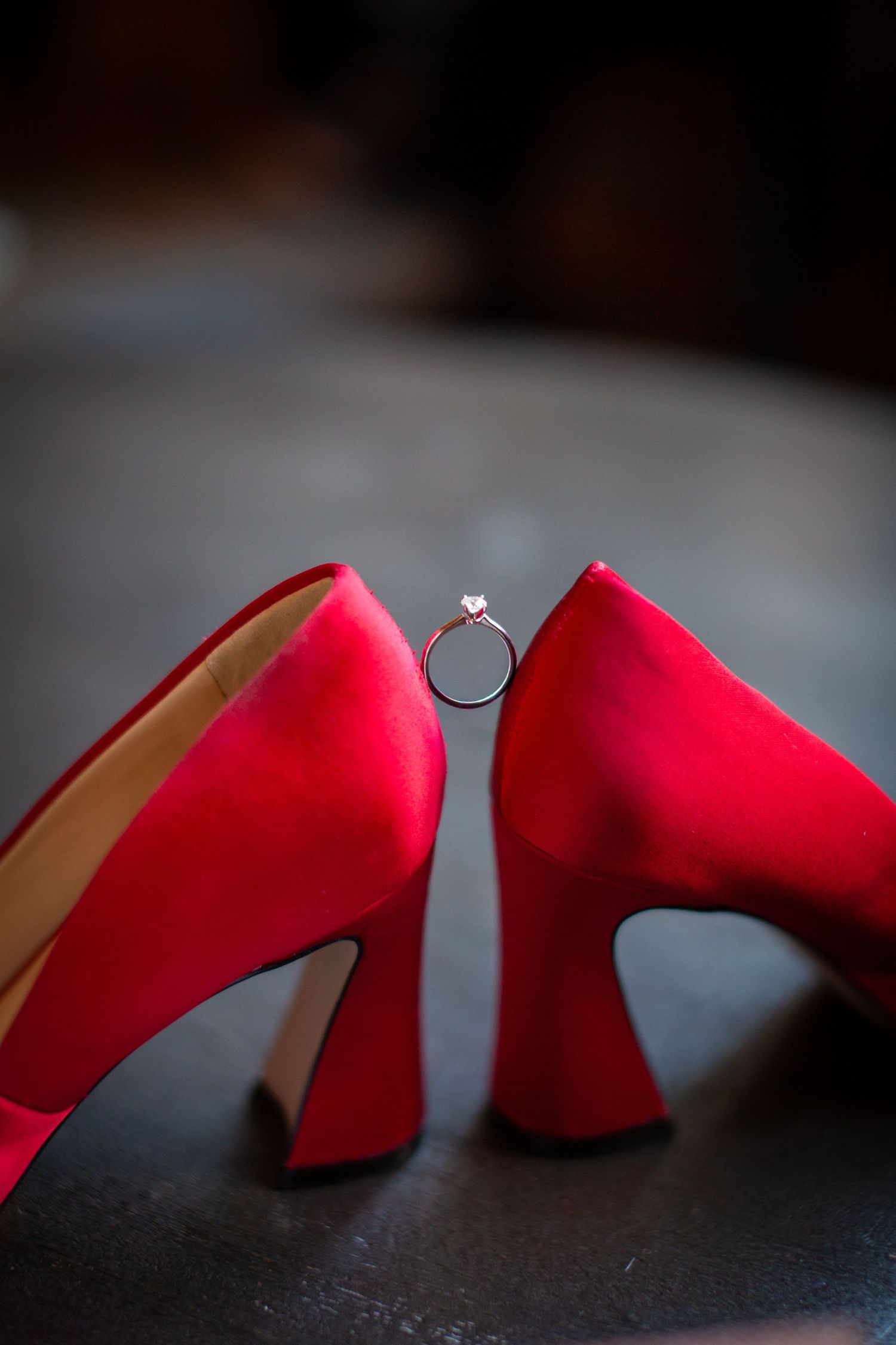Red high heel shoes with engagement ring balanced between them in dramatic lighting.