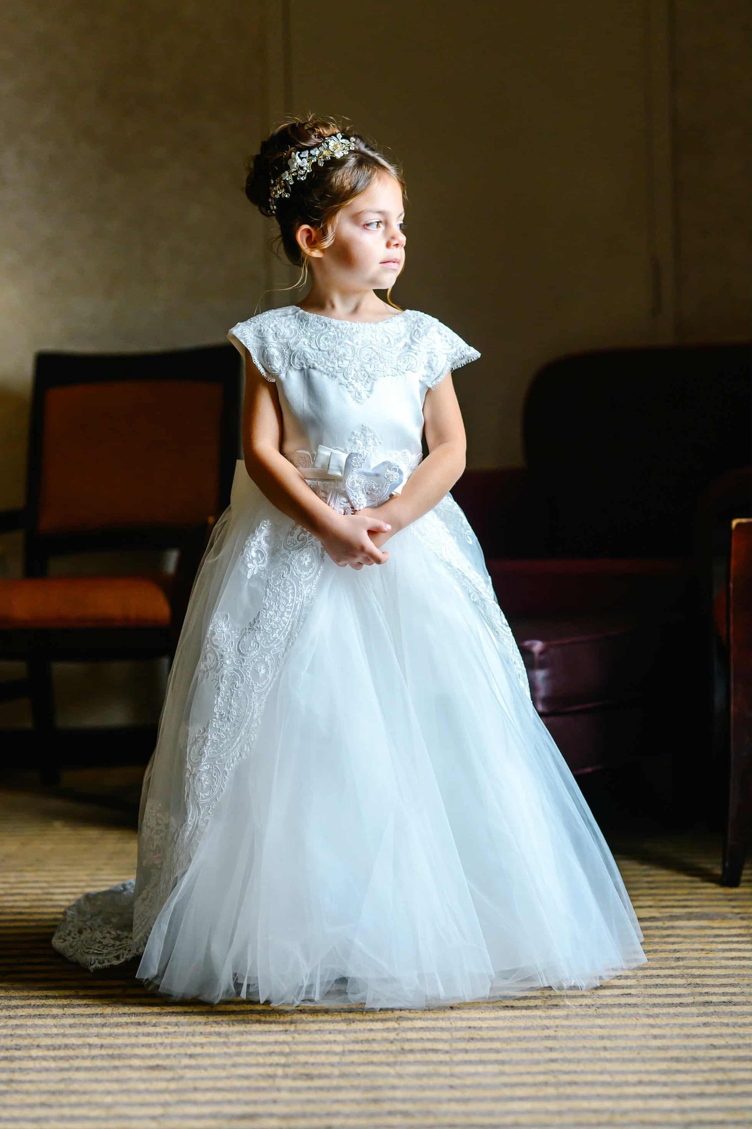 A small child in a flowing white lace and tulle dress poses elegantly in a series of wedding-style portraits.