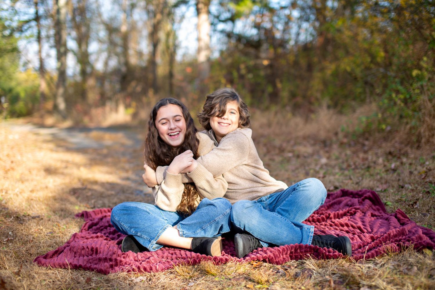 Two people lie on a burgundy blanket in autumn leaves while enjoying a sunny day outdoors.