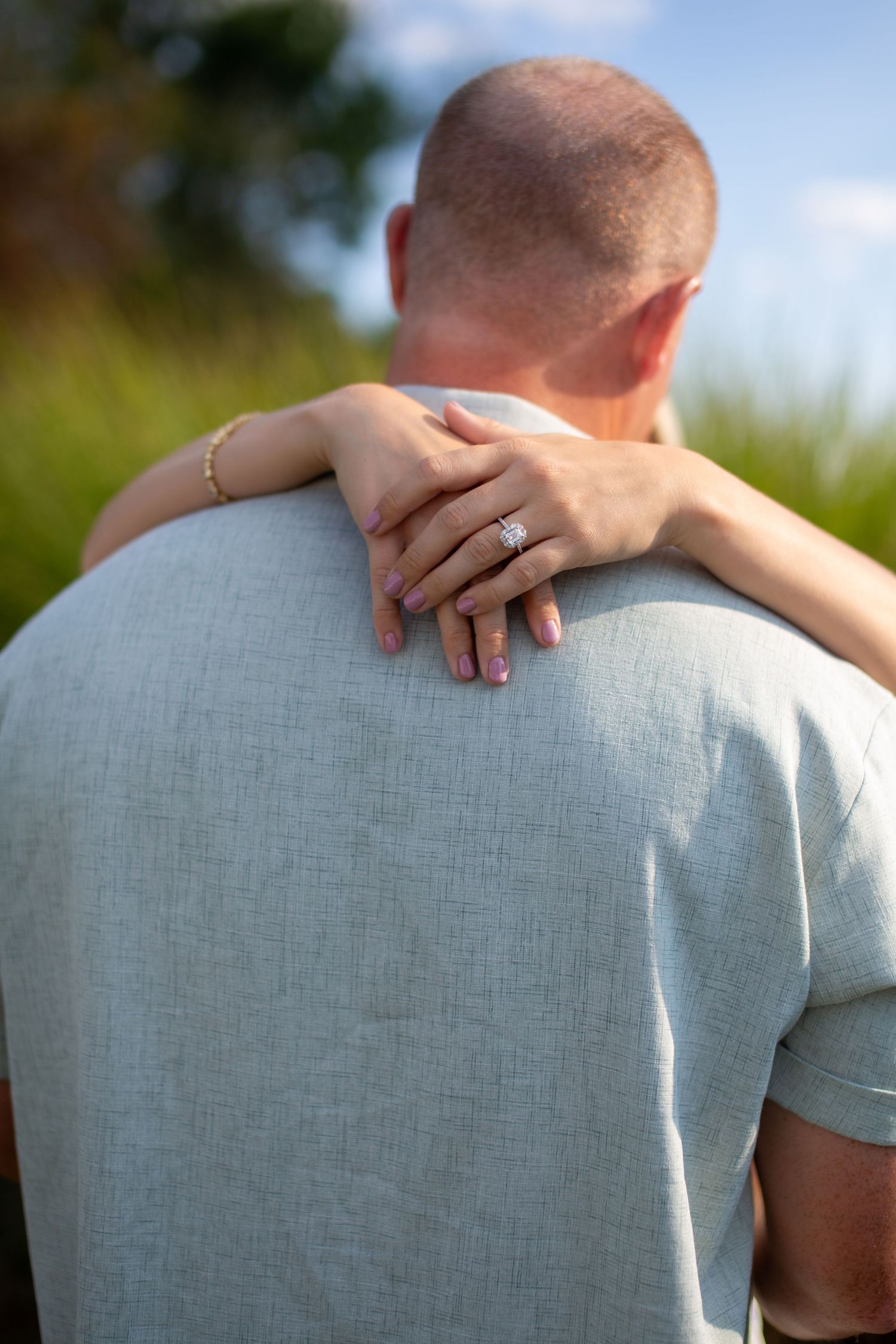Close-up of hands with engagement ring embracing from behind against a summer background.