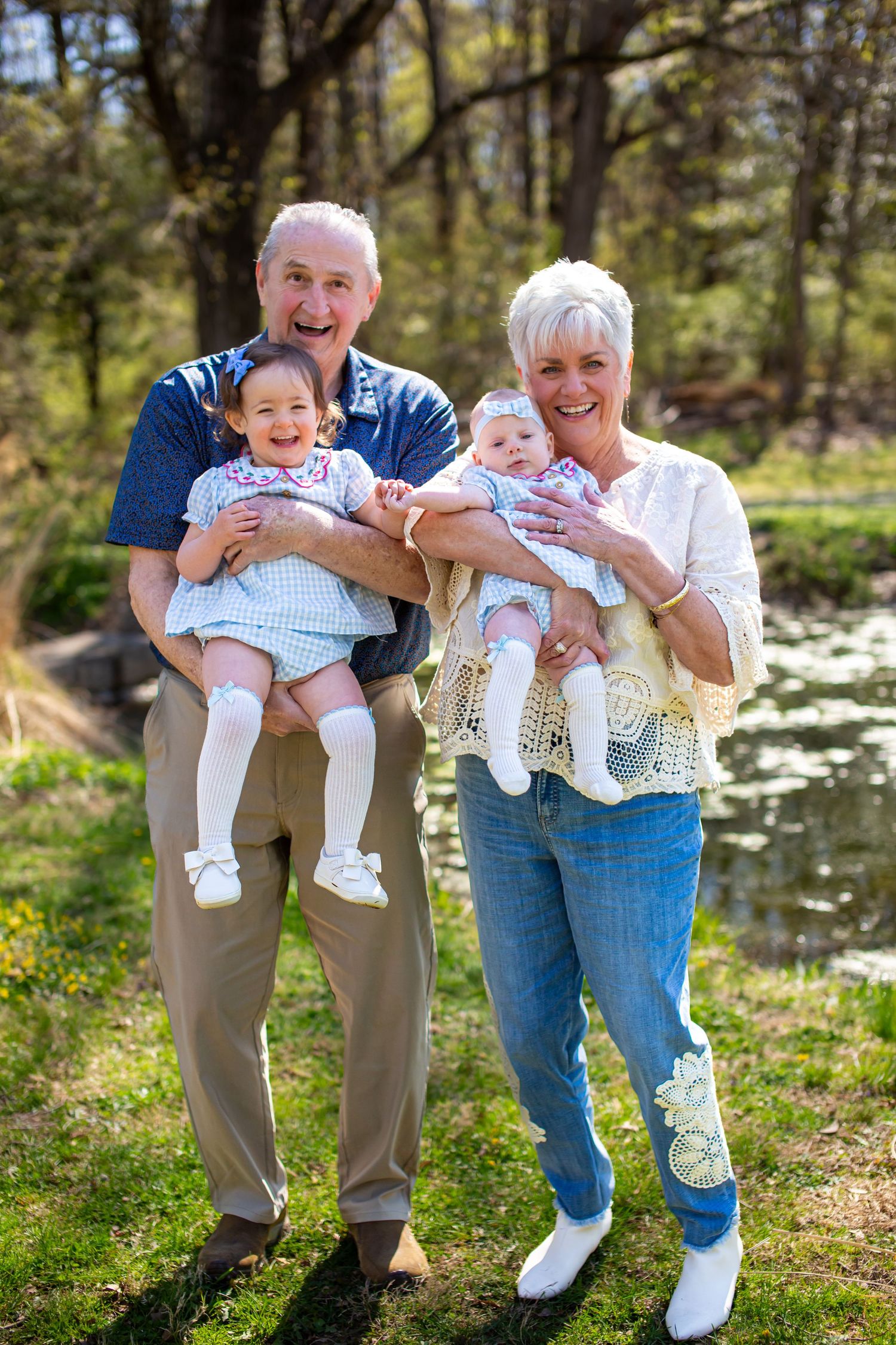 Grandparents smiling outdoors holding two baby girls near a pond in a sunny green park.