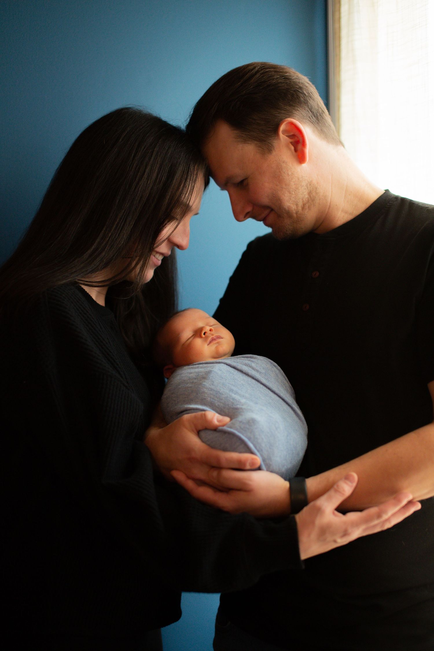 Parents sharing an intimate moment with their swaddled newborn against a blue wall.