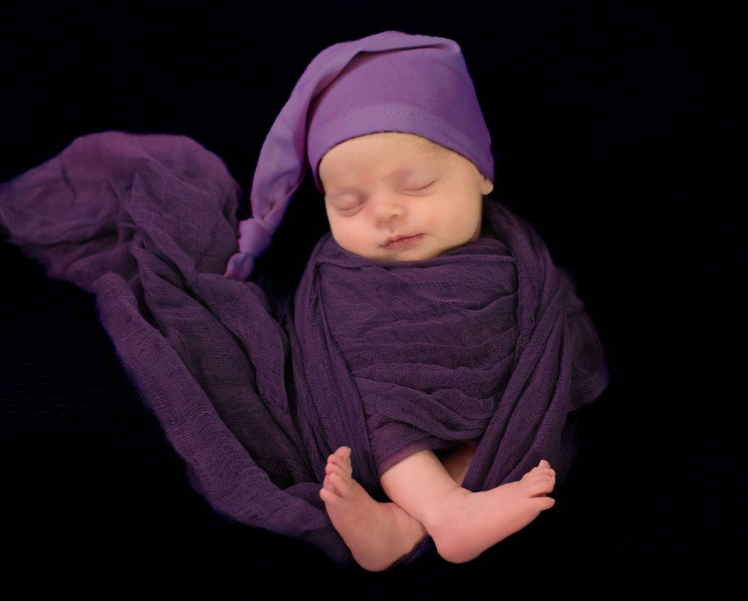 A sleeping newborn wrapped in purple fabric against a black background.