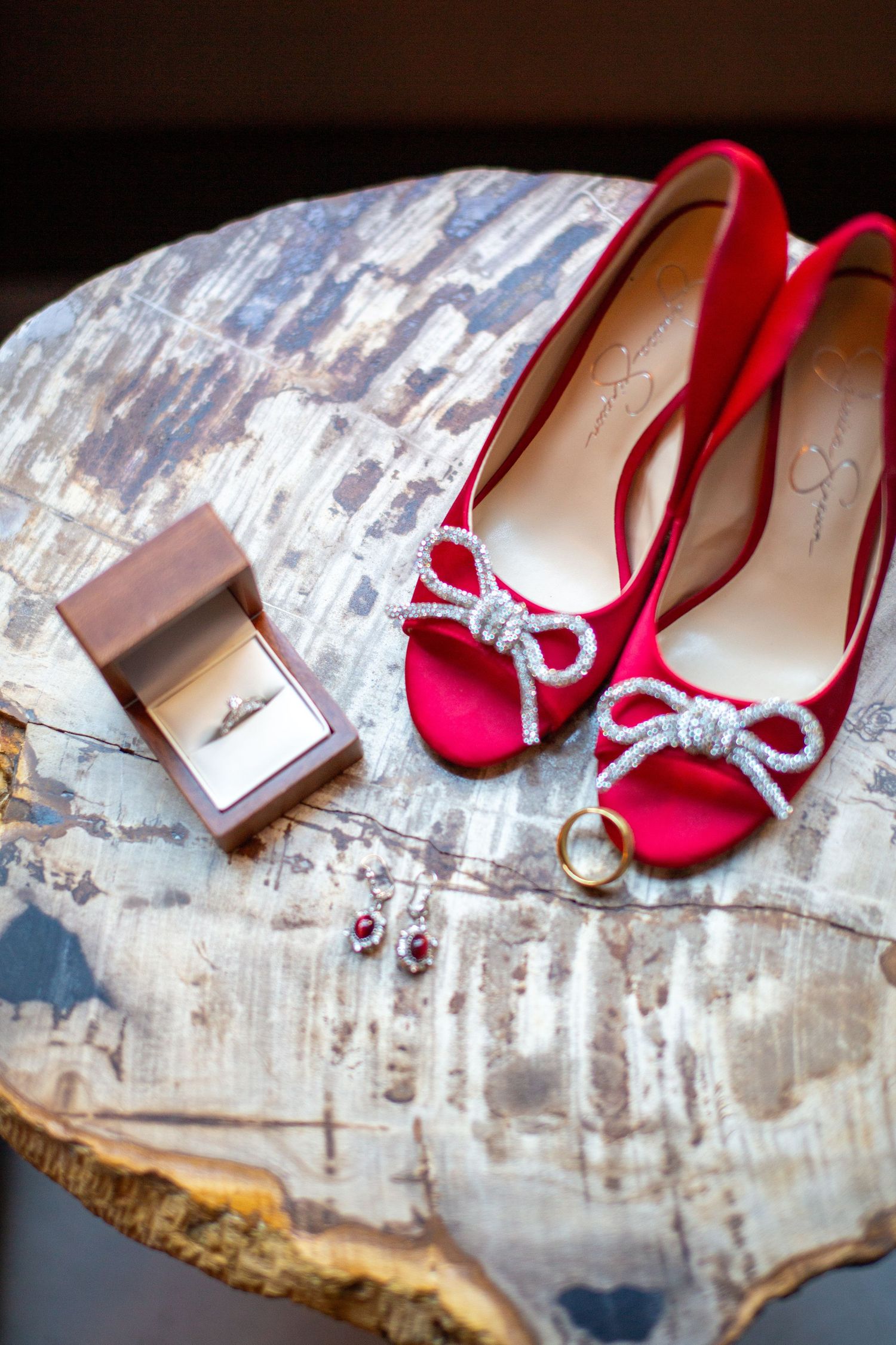 Red bridal shoes with crystal embellishments displayed on a rustic surface with wedding accessories.