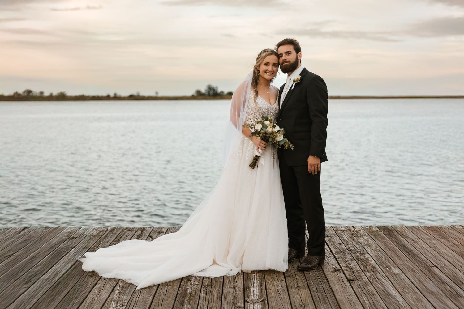 A couple poses on a wooden dock at sunset, with the bride in a flowing white gown holding a bouquet.