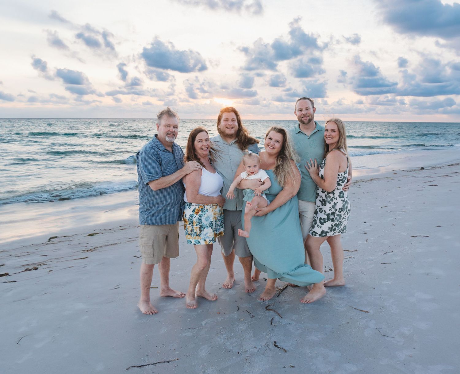 A group stands together on a sandy beach at sunset with waves and cloudy sky in the background.