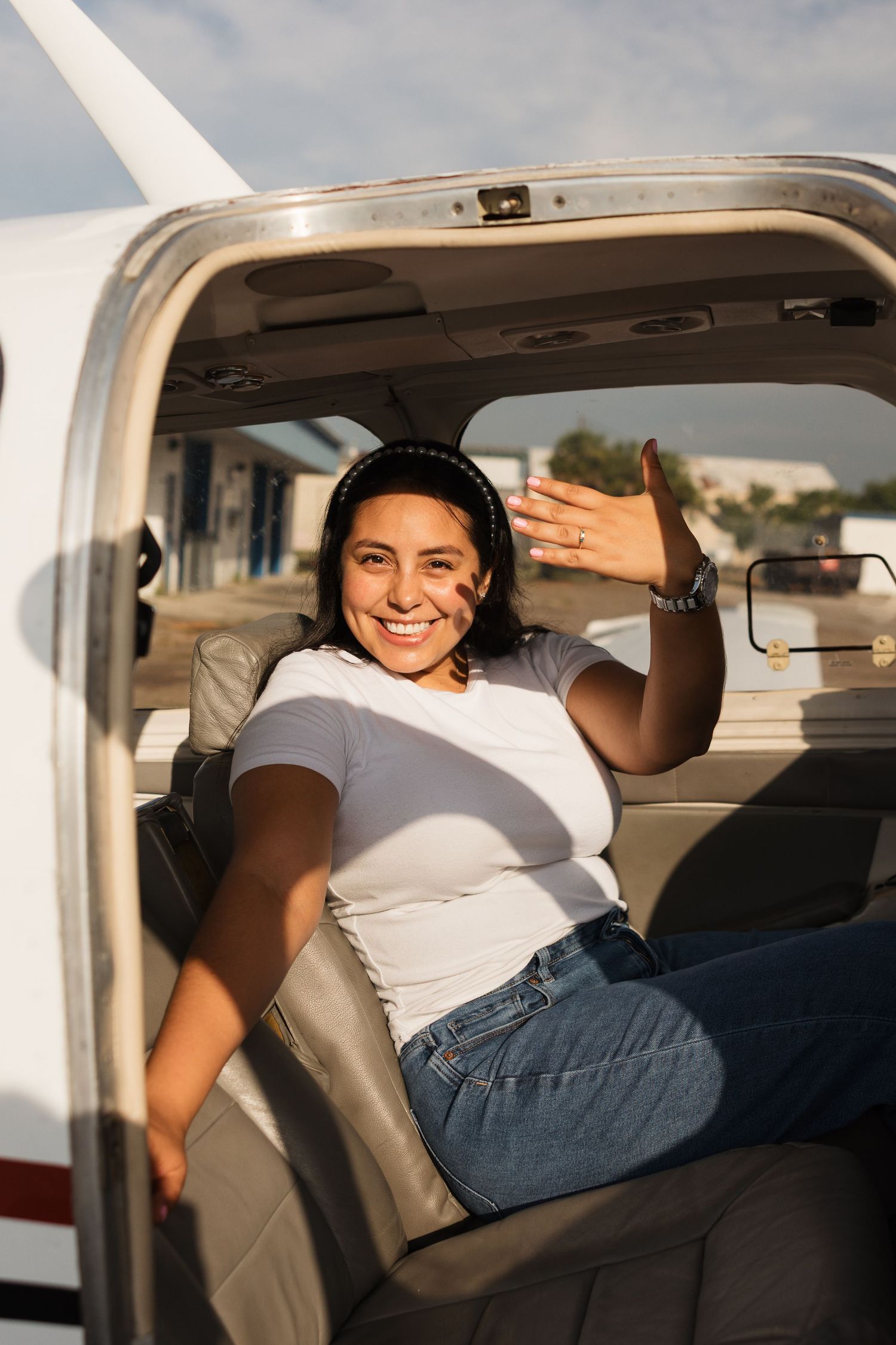 A passenger waves from inside a small private aircraft, wearing a white t-shirt and jeans in warm sunlight.