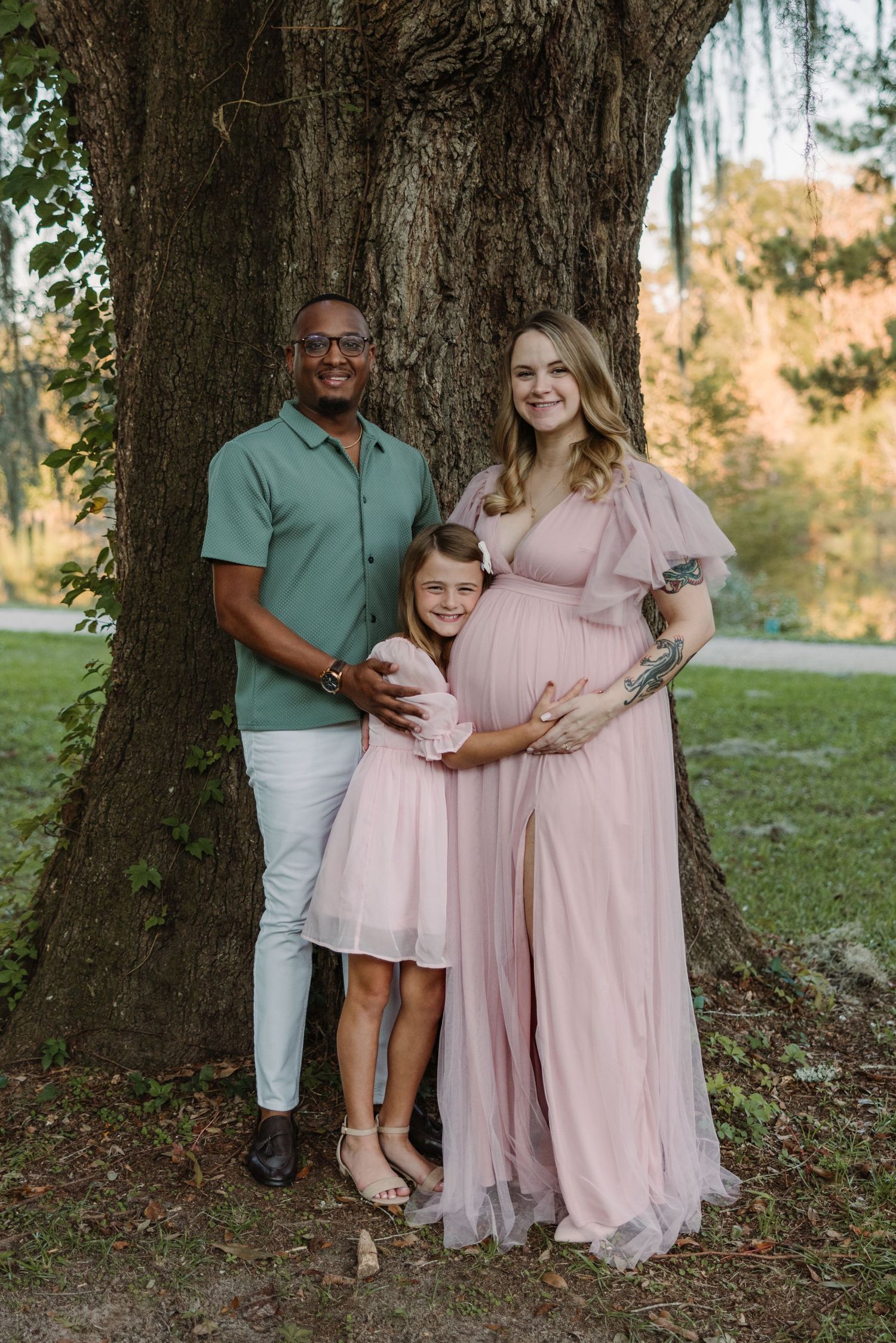 A family poses together under a large tree outdoors, with coordinated pink and mint outfits for a maternity photo session.