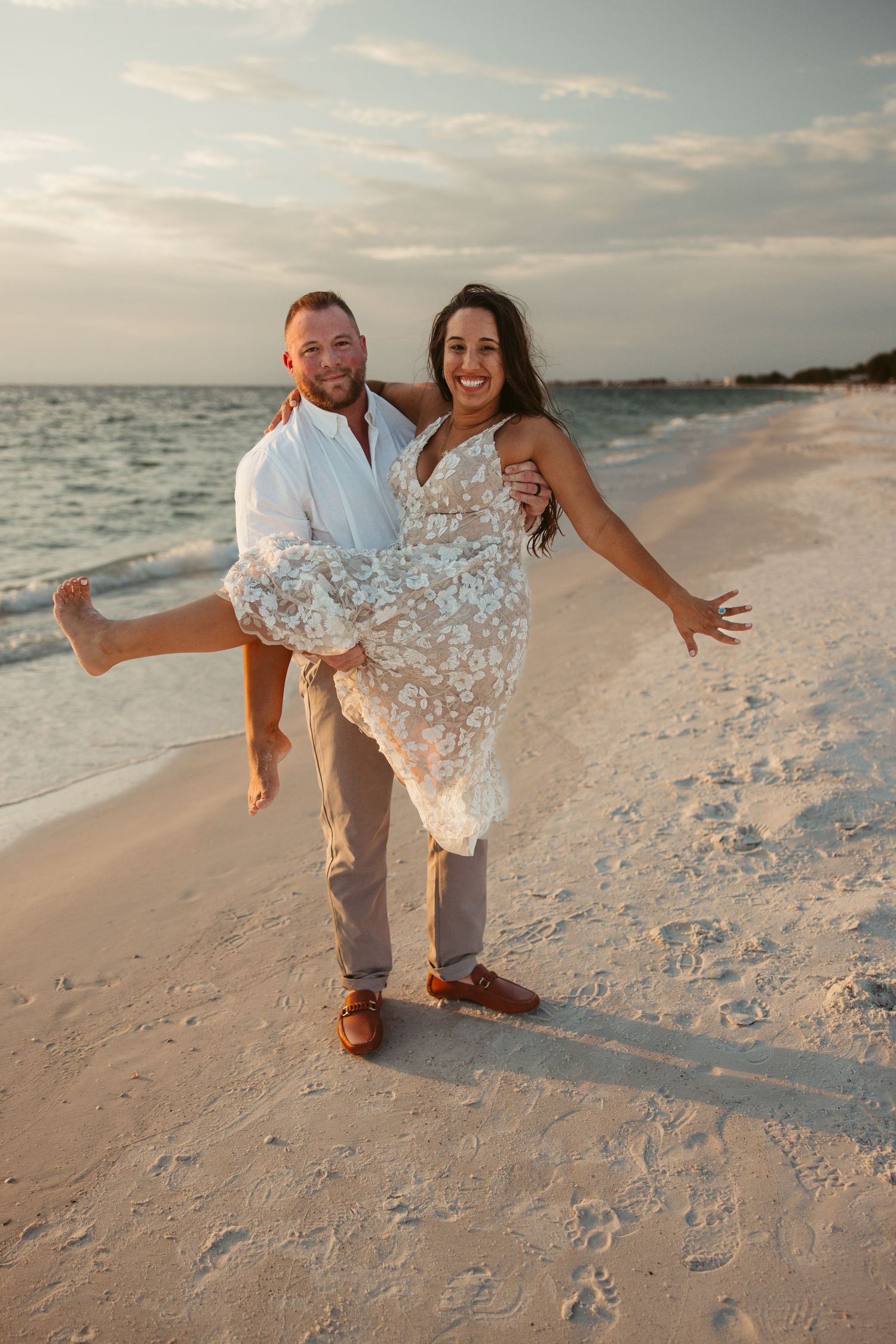A joyful couple poses on a sunset beach, with one partner carrying the other wearing a floral lace dress.