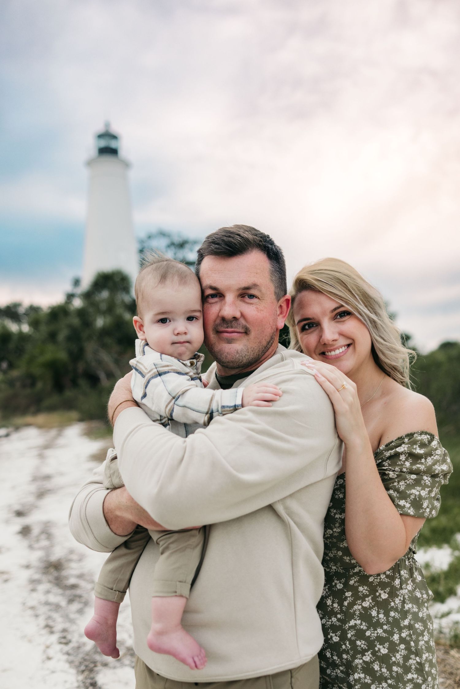 A family poses together on a sandy beach path with a lighthouse visible in the background on a cloudy day.