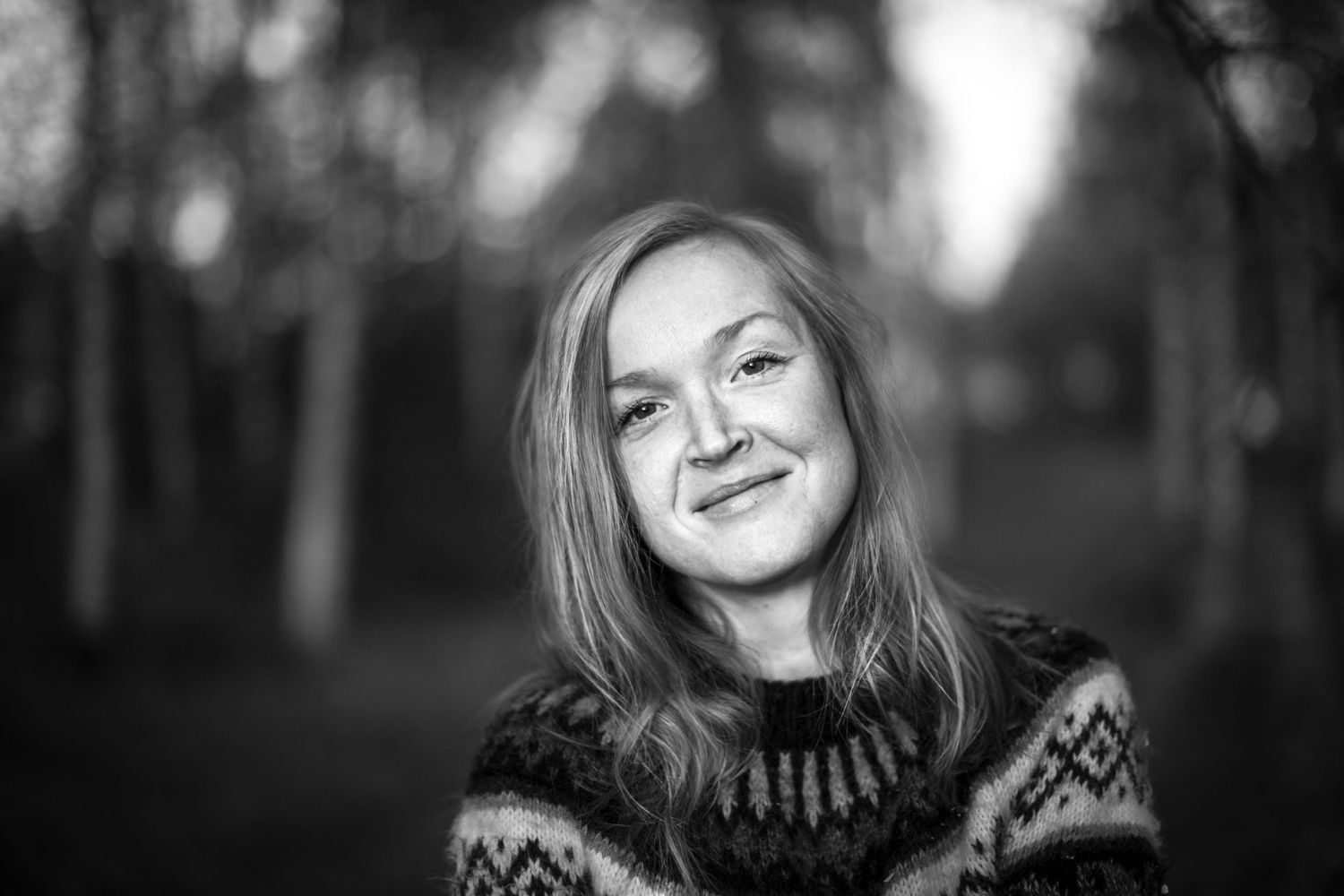 Black and white close-up portrait of smiling woman in patterned sweater with blurred autumn trees background.