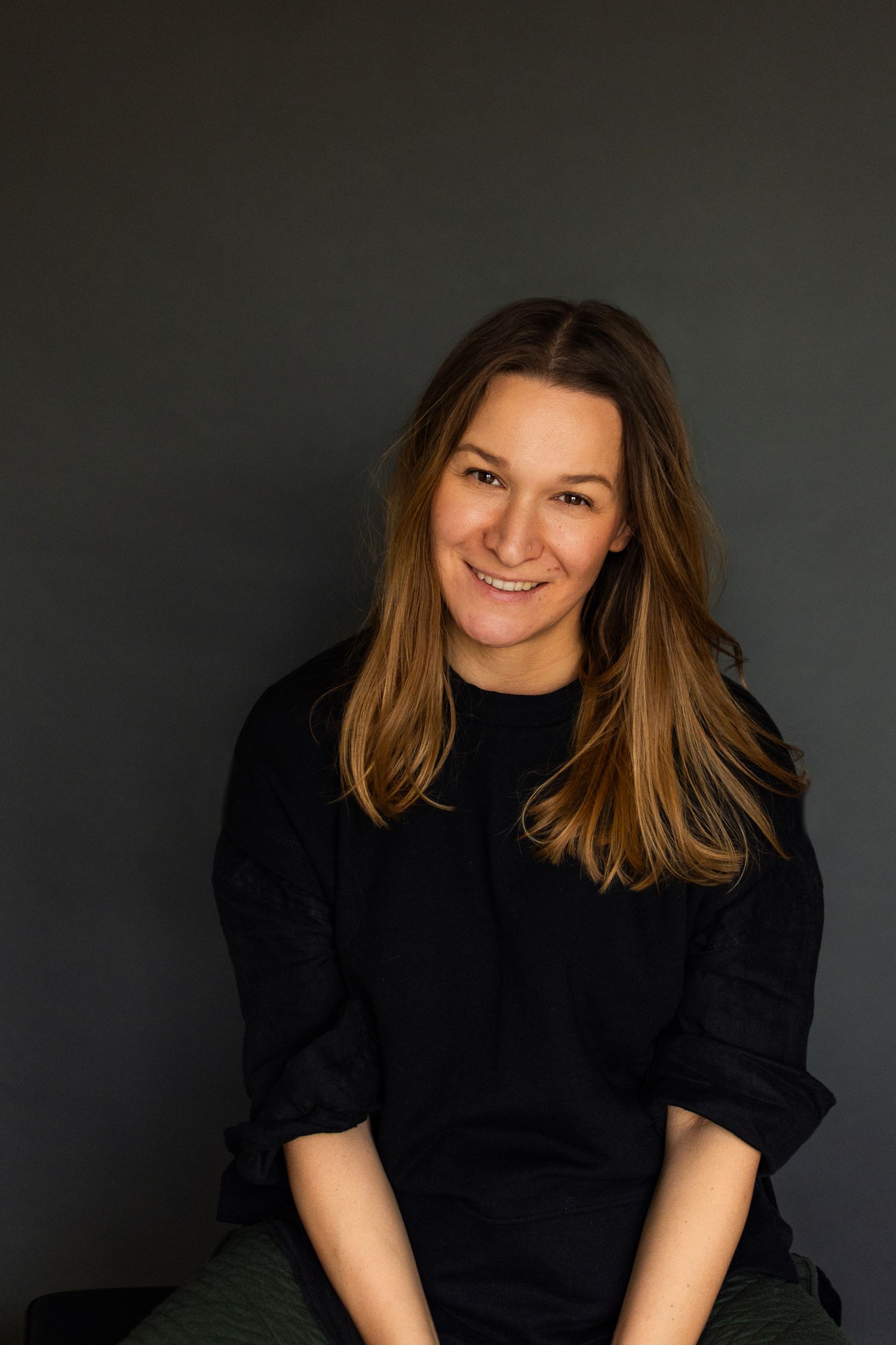 Smiling woman with long brown hair wearing a black top, seated against a dark grey background. Cambridge, UK