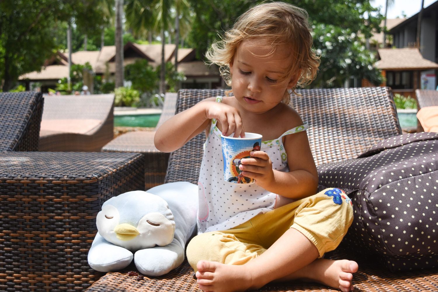 Toddler girl sitting on wicker chair eating from blue cup beside penguin stuffed animal outdoors.