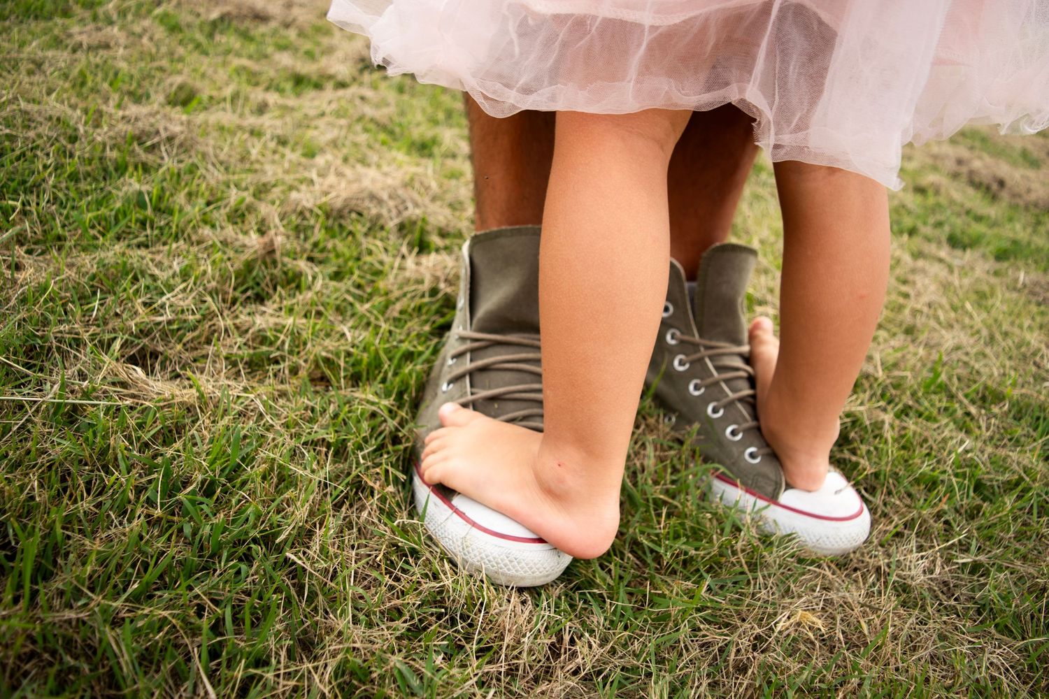 A little barefoot child stands on the father's feet on the green grass in the Meadows, Cambridge, UK