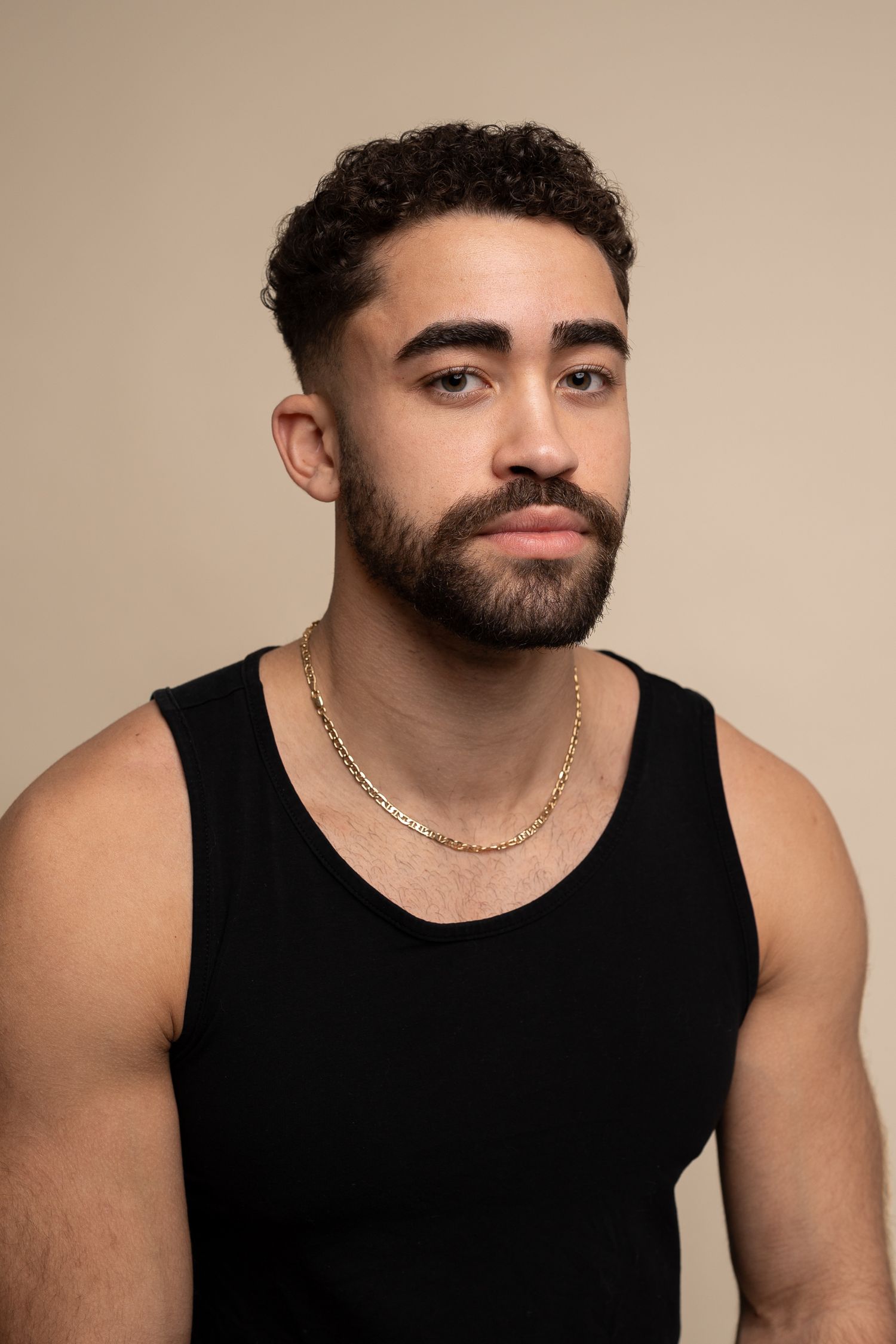 A headshot of a bearded young man wearing a black tank top with a gold chain necklace against a beige background, Studio in Cambridge, UK