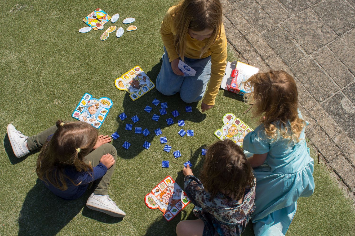 Four children sitting on grass playing a board game with blue tokens outdoors on a sunny day.