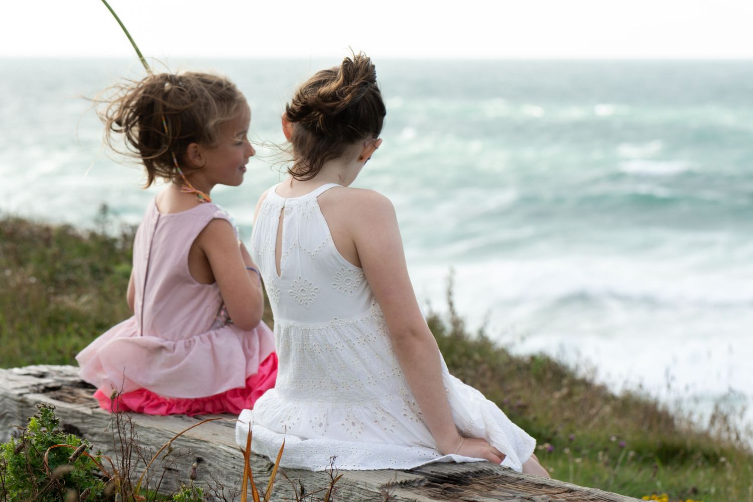 Two figures sit together on a rocky cliff overlooking the ocean on a breezy day.